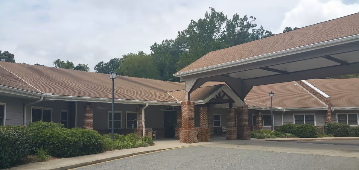 Exterior view of a single-story building with a covered entrance supported by brick columns. The building has a brown shingled roof and gray siding, surrounded by green bushes and trees under a cloudy sky.