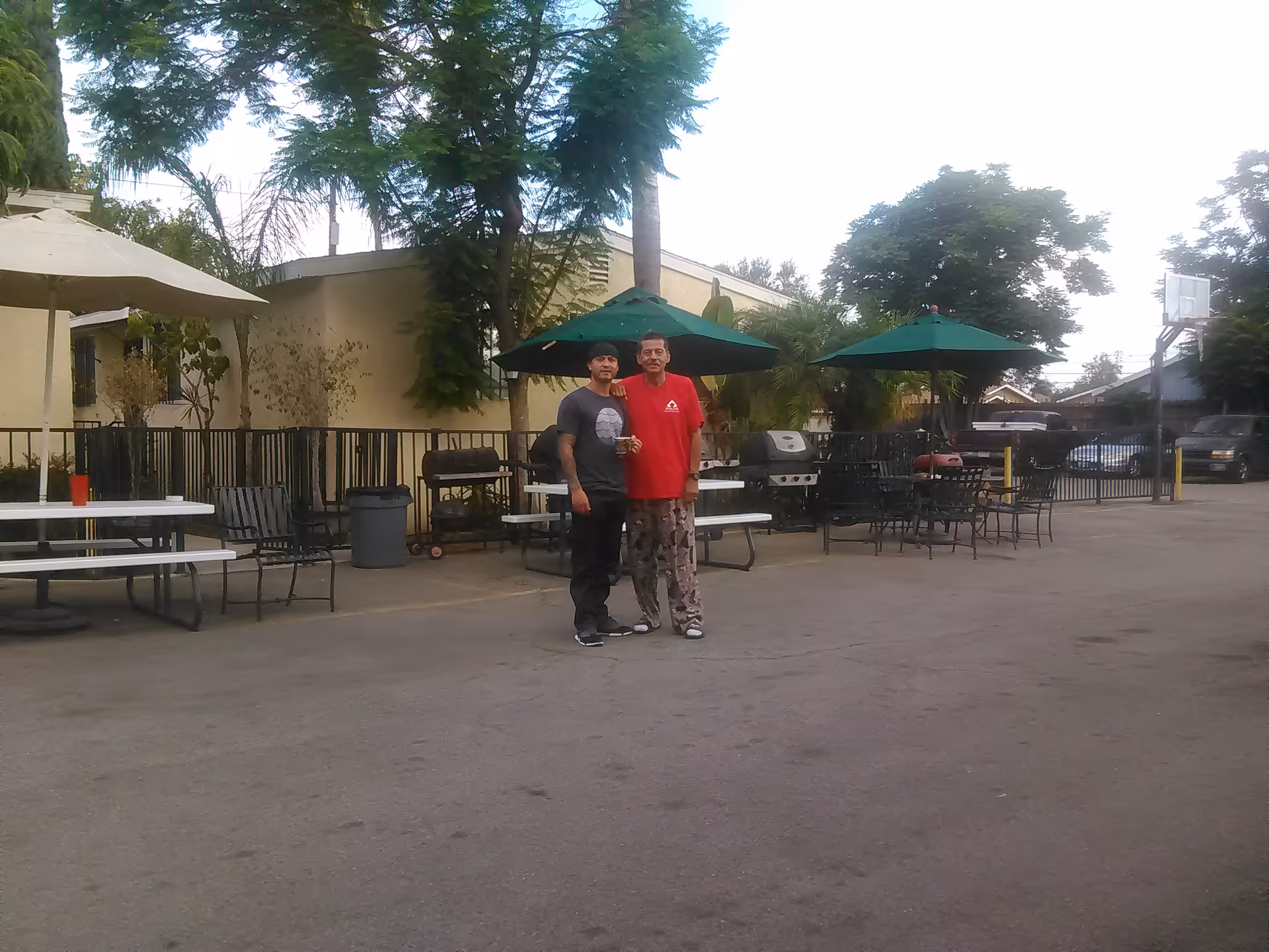 Two men stand in an outdoor seating area with green umbrellas and picnic tables.