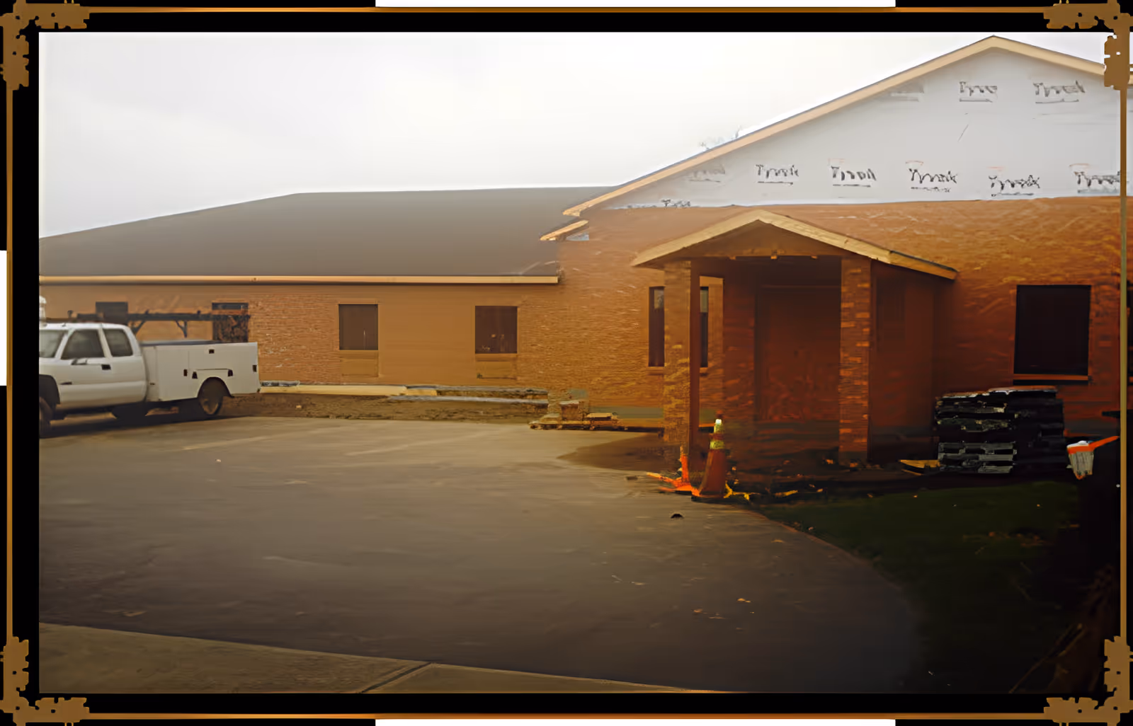 Front exterior of a brick senior living building under construction with a parking lot, a white utility truck, entry portico, traffic cones and stacks of construction materials.