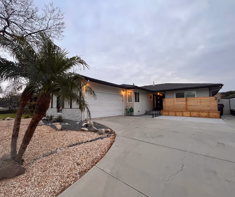 Single-story Pioneer Care Home exterior with driveway, palm trees, and wooden privacy screen.
