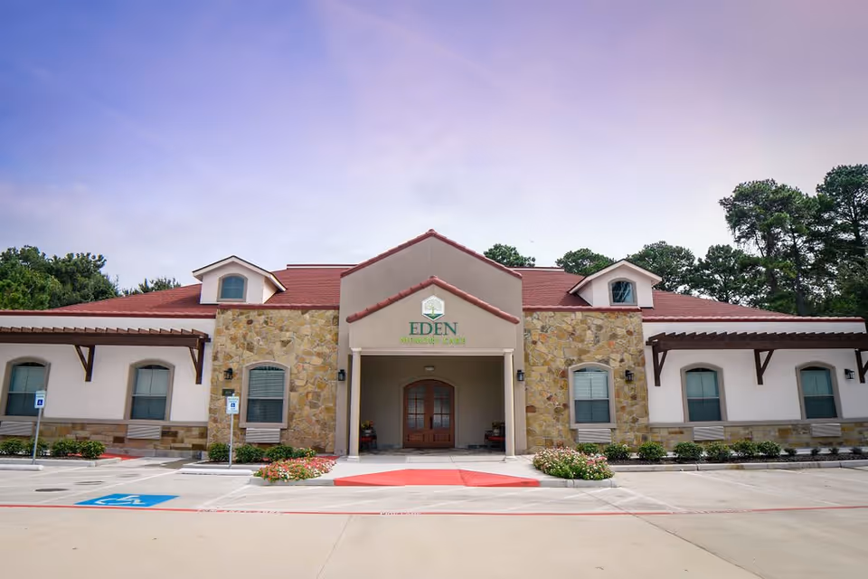 Front exterior view of a single-story building with stone and stucco facade, red roof, and a covered entrance. The building has multiple windows with white blinds and is surrounded by a parking lot with handicap parking spaces and landscaped flower beds. Trees are visible in the background under a partly cloudy sky.