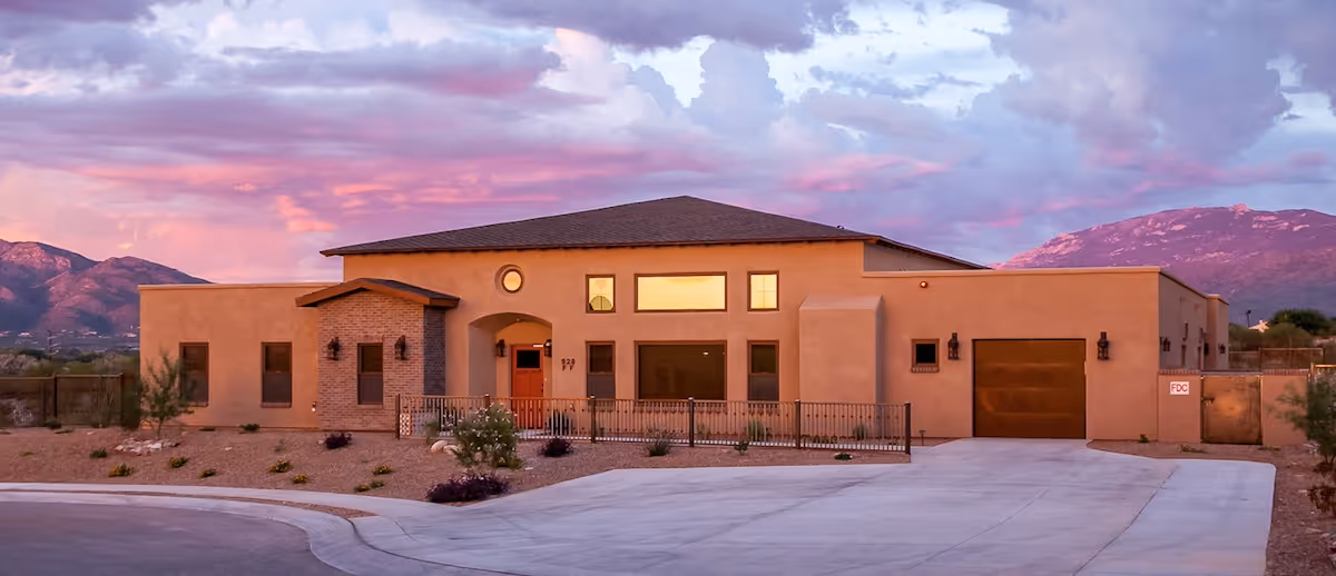 Exterior view of a single-story building with a southwestern architectural style, featuring a flat roof, beige stucco walls, and a brick accent around the entrance. The building is set against a backdrop of mountains and a colorful sky at sunset. There is a driveway and some desert landscaping in front.