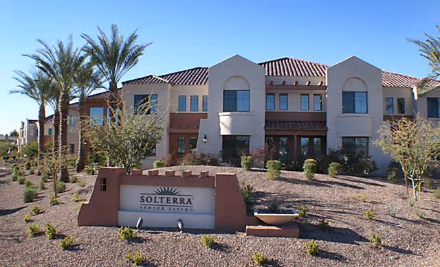 Exterior view of Solterra Senior Living facility with a two-story building featuring beige and brown stucco walls, red tile roof, and several windows. The foreground shows desert landscaping with small bushes, palm trees, and a sign that reads 'Solterra Senior Living'.