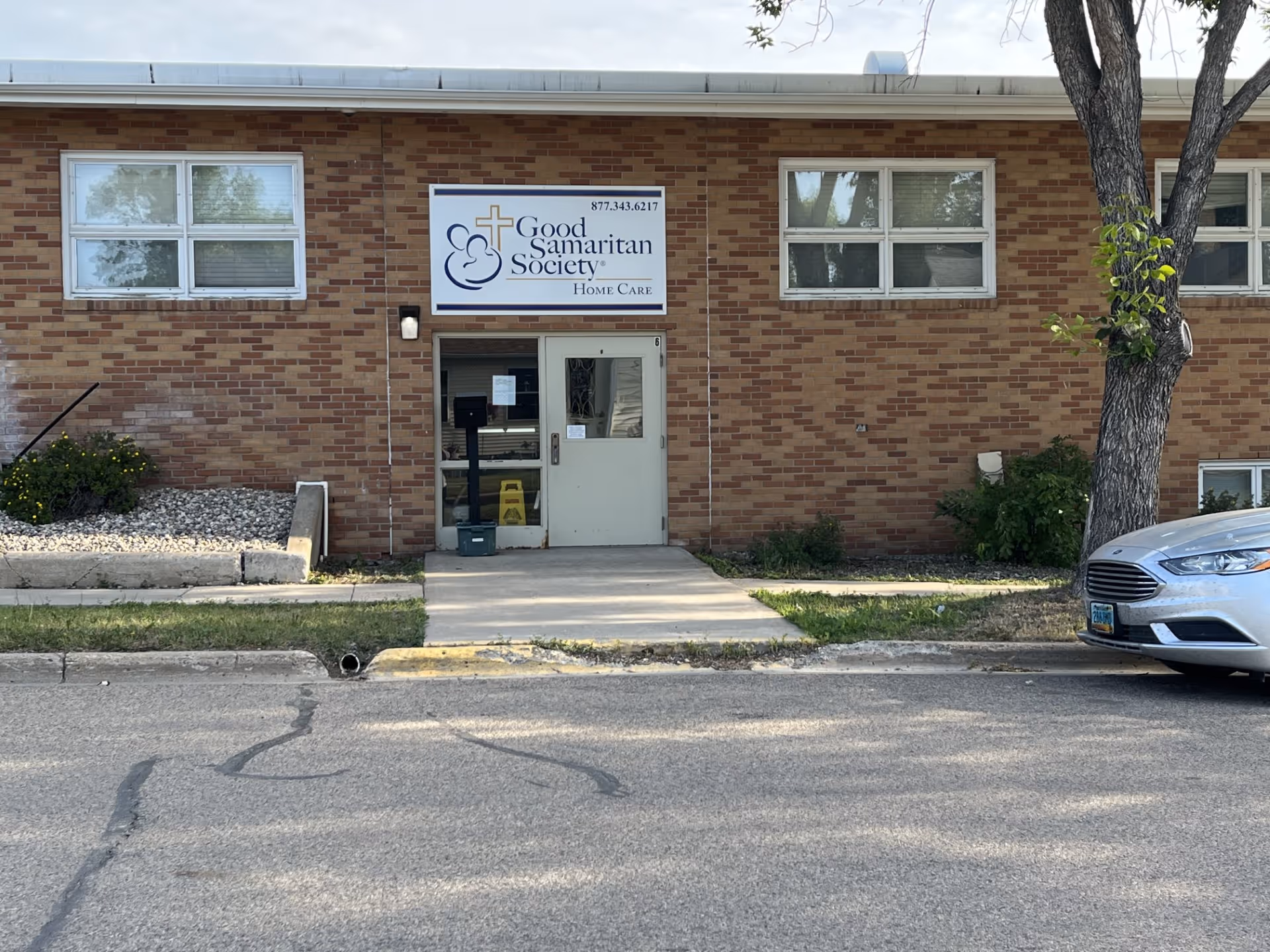 Brick building entrance of Good Samaritan Society Home Care with sign and parked car.