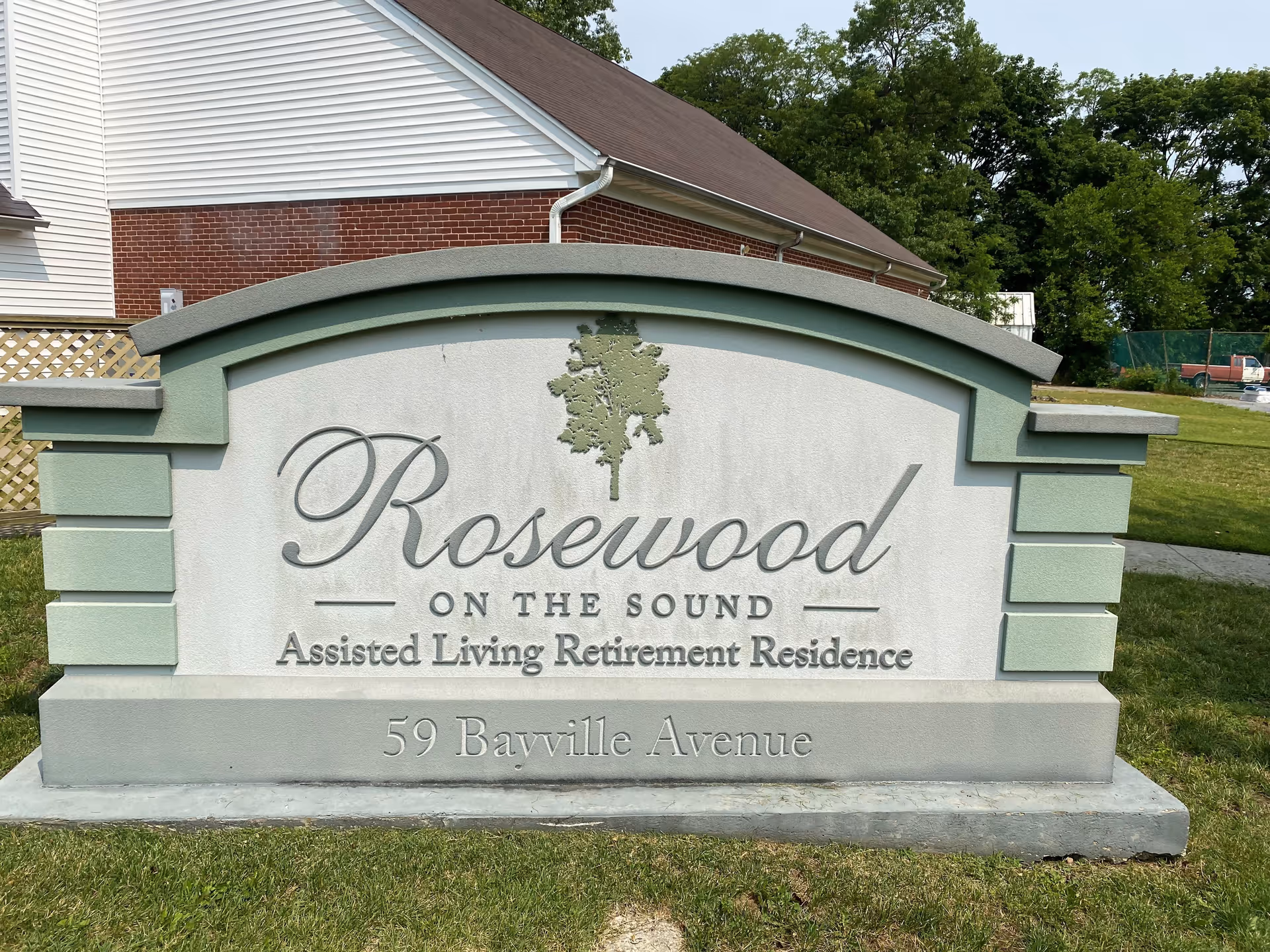 A large outdoor sign for Rosewood On The Sound Assisted Living Retirement Residence, located at 59 Bayville Avenue. The sign is made of stone with green and white colors, featuring a tree logo above the facility name. Behind the sign, there is a building with white siding and red brick, and green trees in the background.