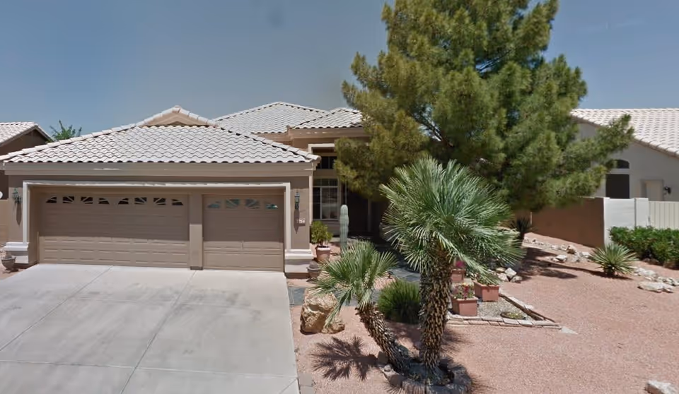 Front exterior view of a single-story house with a three-car garage, beige tiled roof, and desert landscaping including palm trees, rocks, and gravel under a clear blue sky.
