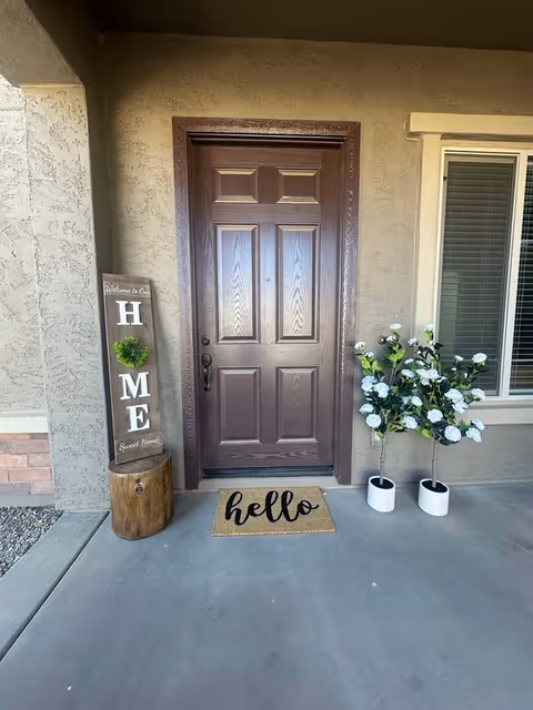 Exterior front entrance with brown door, welcome sign, hello mat, and potted white flowers.