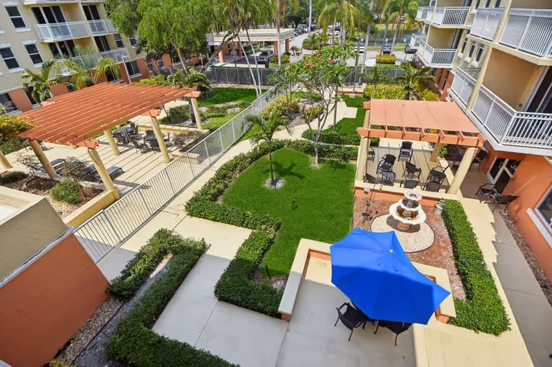 Aerial view of a landscaped courtyard between apartment buildings with pergolas, seating areas, a fountain and a blue umbrella.