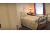 A small bedroom in a senior living facility with a bed, nightstand, dresser, and window.