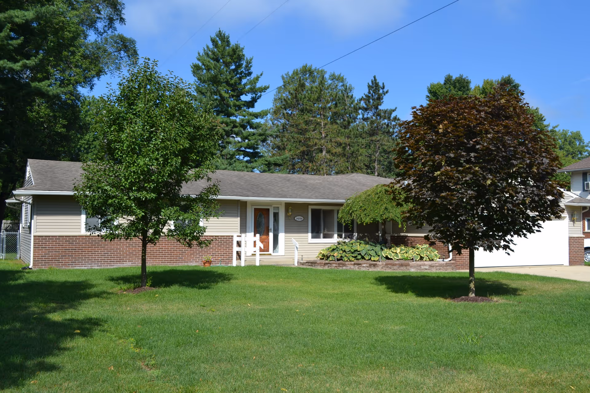 Single-story ranch-style house with a green front lawn, two trees, and a driveway under a blue sky.