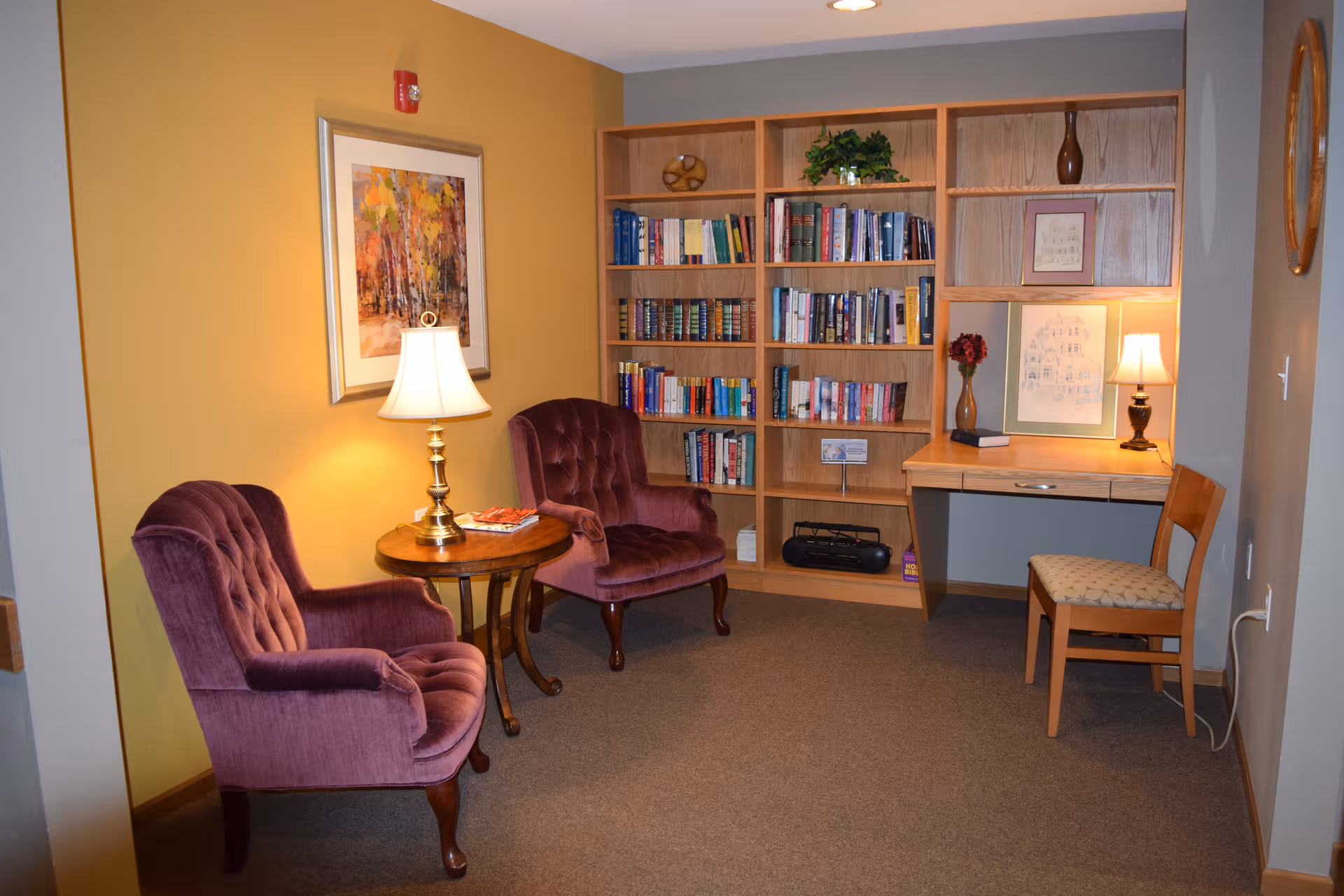 Cozy seating area in senior living facility with two purple chairs, bookshelf, desk, lamps.