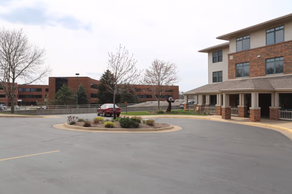 Exterior view of Stonehill Health Center showing the front entrance with a covered porch supported by brick columns, a circular driveway with a landscaped island in the center, a red car parked near the entrance, leafless trees, and a multi-story brick building in the background under a cloudy sky.