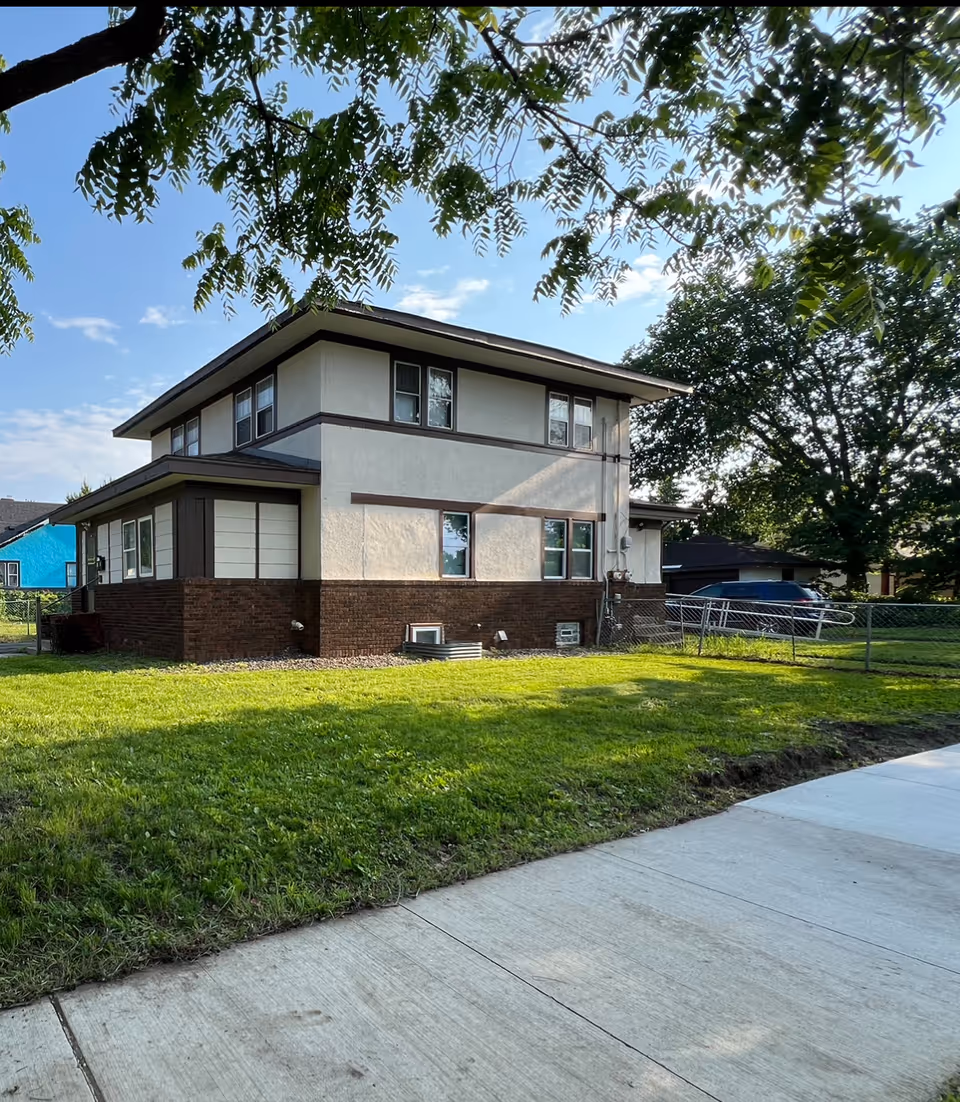 Two-story residential building with brick base, white upper walls, trees, and a grassy yard.