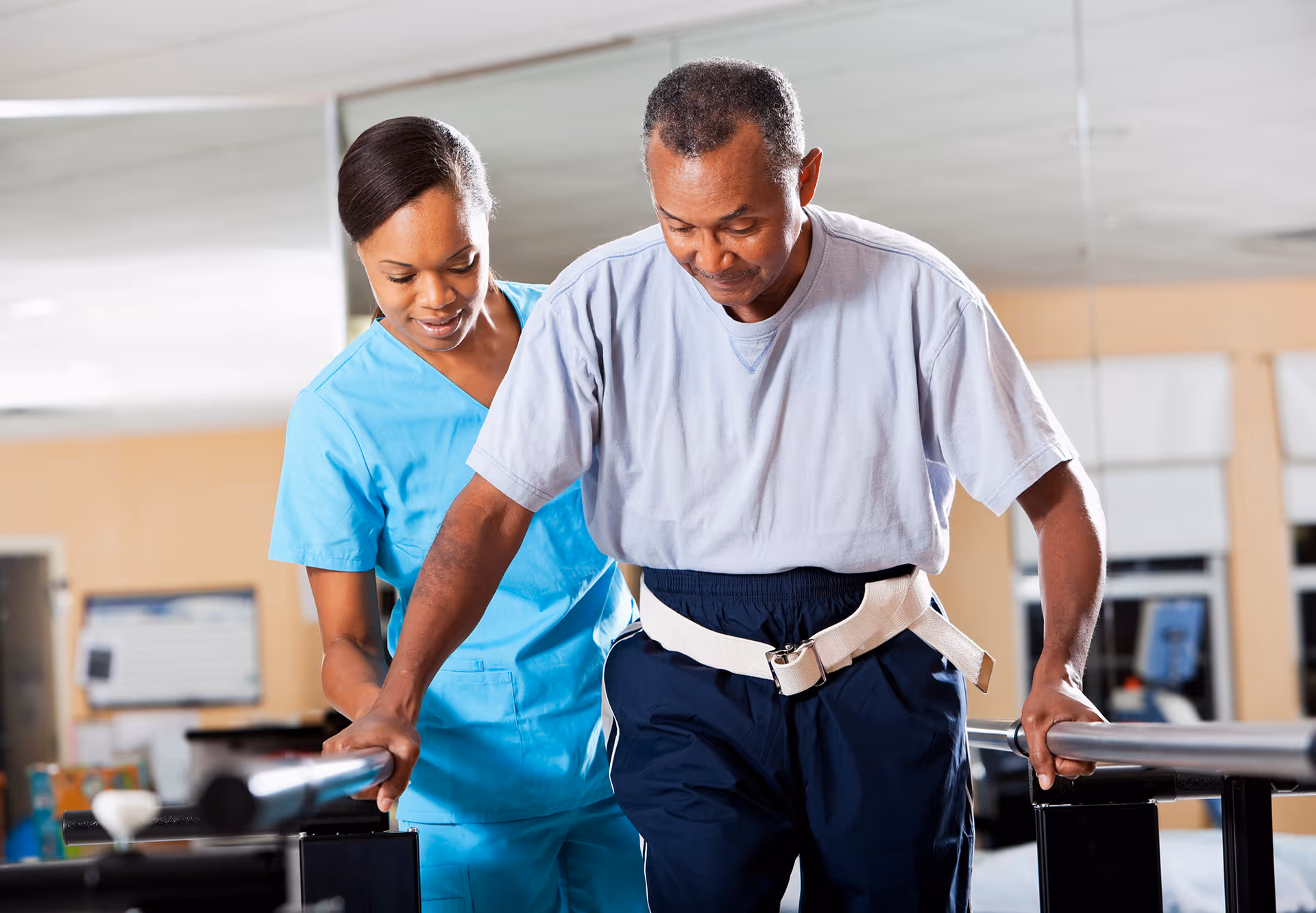A healthcare worker in blue scrubs assists an elderly man using parallel bars for walking practice in a rehabilitation or therapy setting.