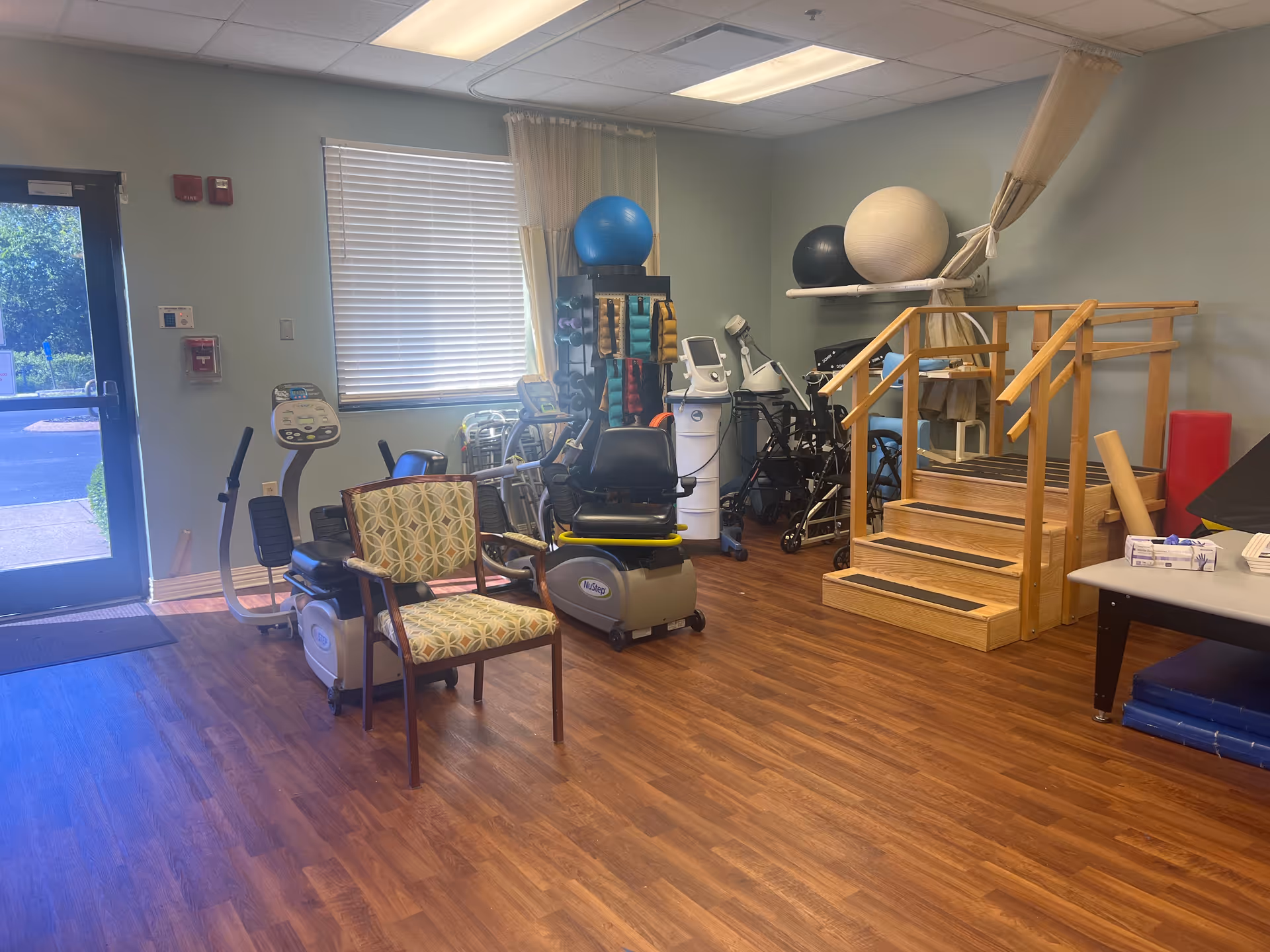 Bright rehab/exercise room with equipment, balance balls, and a wooden staircase.