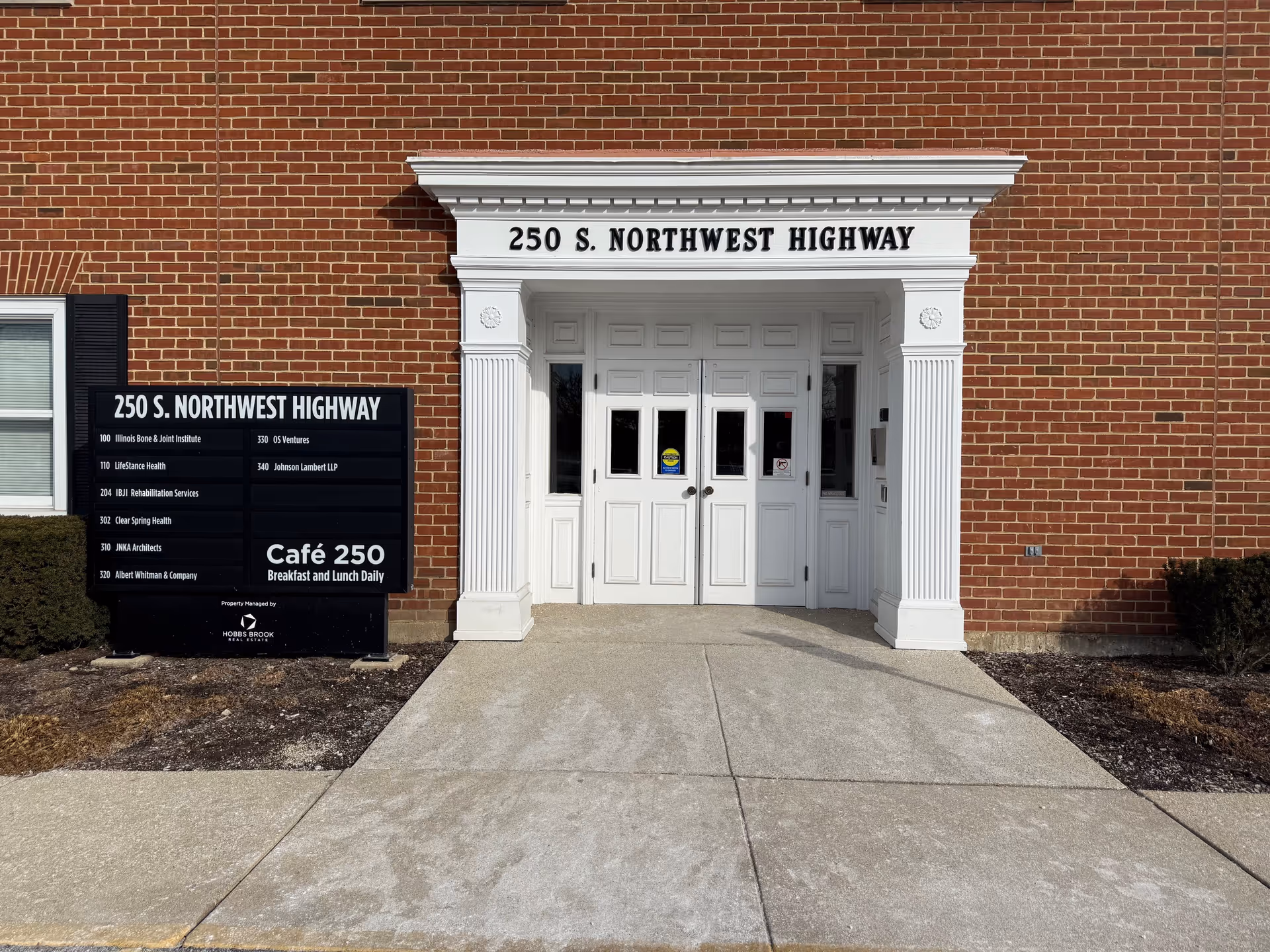 Brick building entrance with white double doors, decorative columns, and a black directory sign.