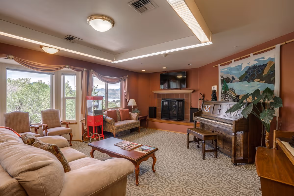 A cozy living room with a beige sofa, two armchairs, a wooden coffee table with magazines, a red popcorn machine, a loveseat, a fireplace with a TV mounted above it, and a wooden piano with a bench. The room has large windows with draped curtains showing a view of trees and hills outside. There is a decorative tapestry hanging on the wall above the piano and a potted plant next to it.