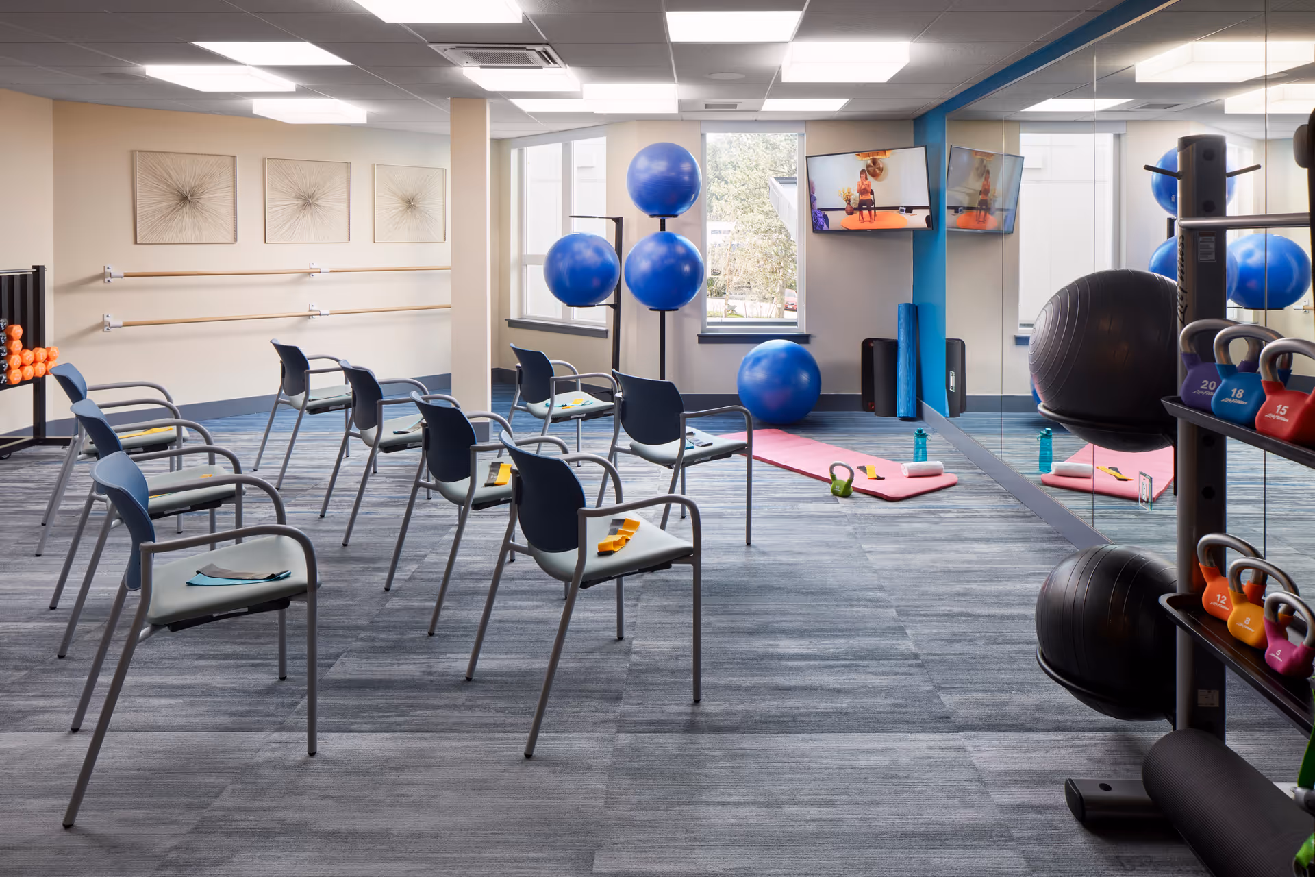 Bright fitness/exercise room with chairs arranged for a class, exercise balls, mats, kettlebells and a mirrored wall.