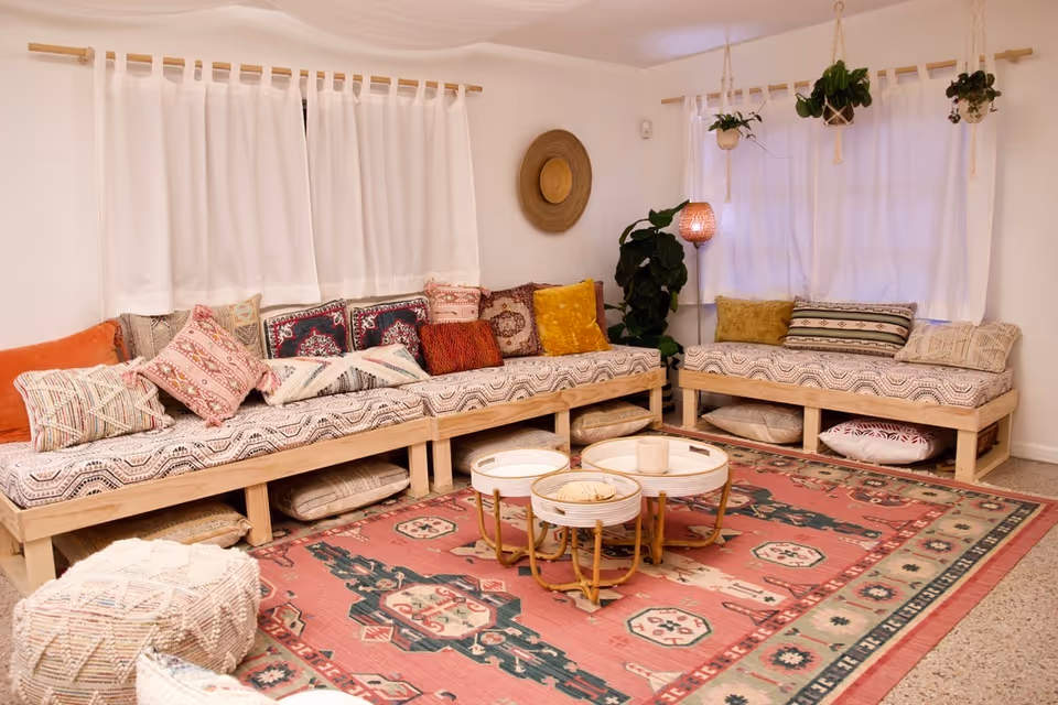 Cozy communal living room with low wooden sofas, colorful cushions, plants, and a patterned rug.