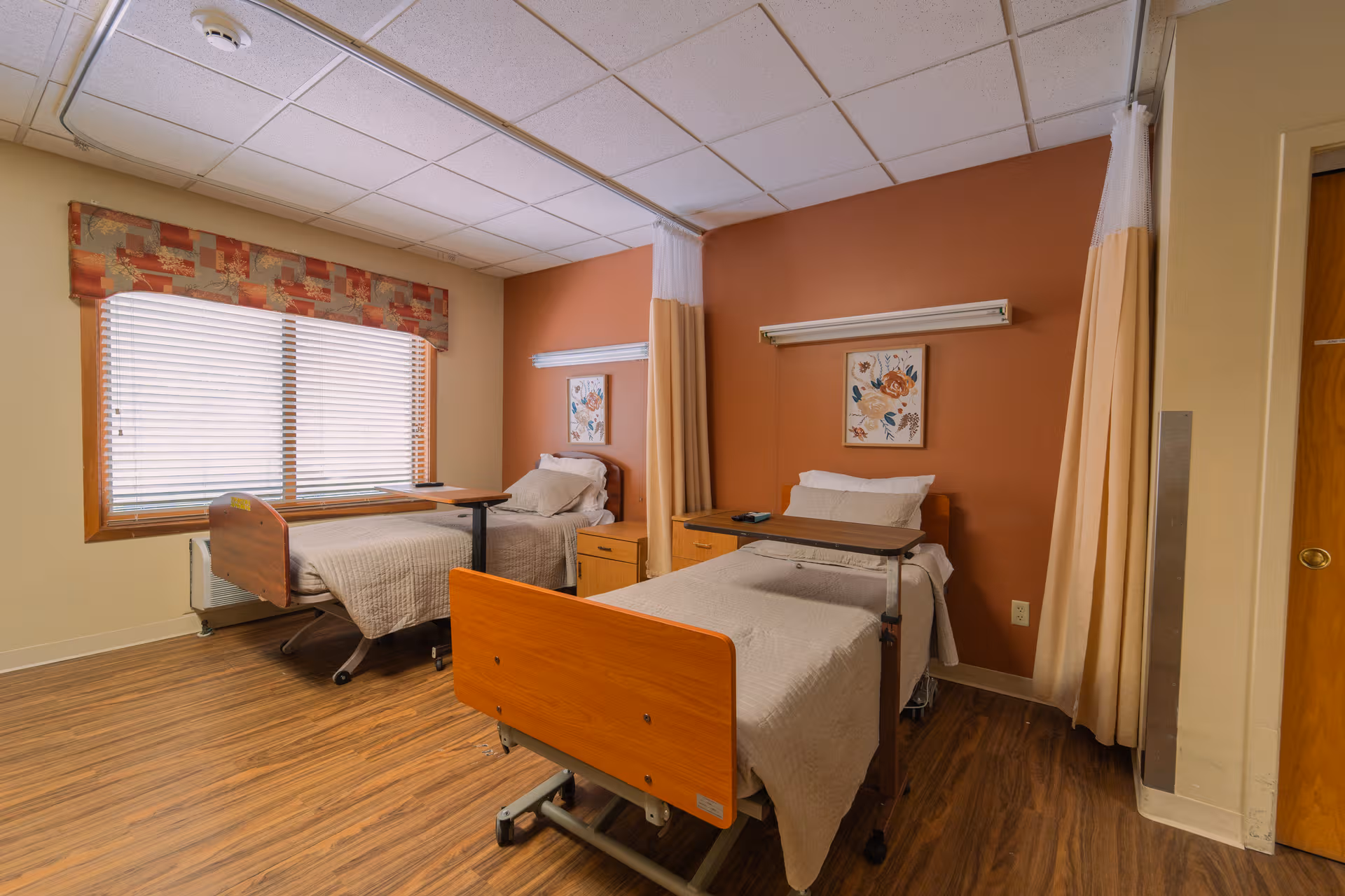 Two hospital-style beds in a shared senior living bedroom with curtains and wood flooring.