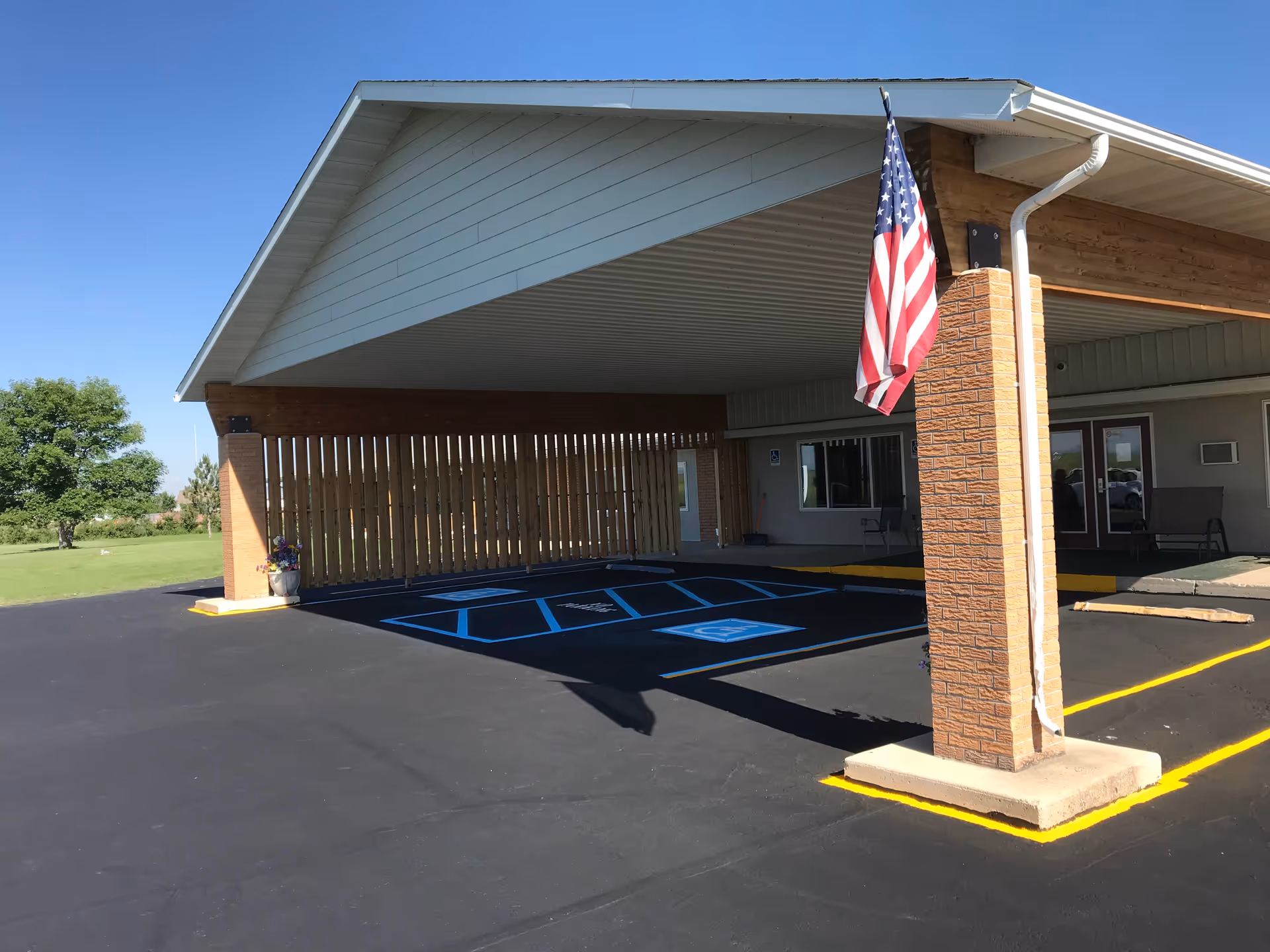 Covered entrance area of a building with an American flag hanging from a brick pillar. There are two handicap parking spaces marked in blue on the asphalt in front of the entrance. The building has a light-colored roof and walls, and there is a wooden fence on one side. A green lawn and trees are visible in the background under a clear blue sky.