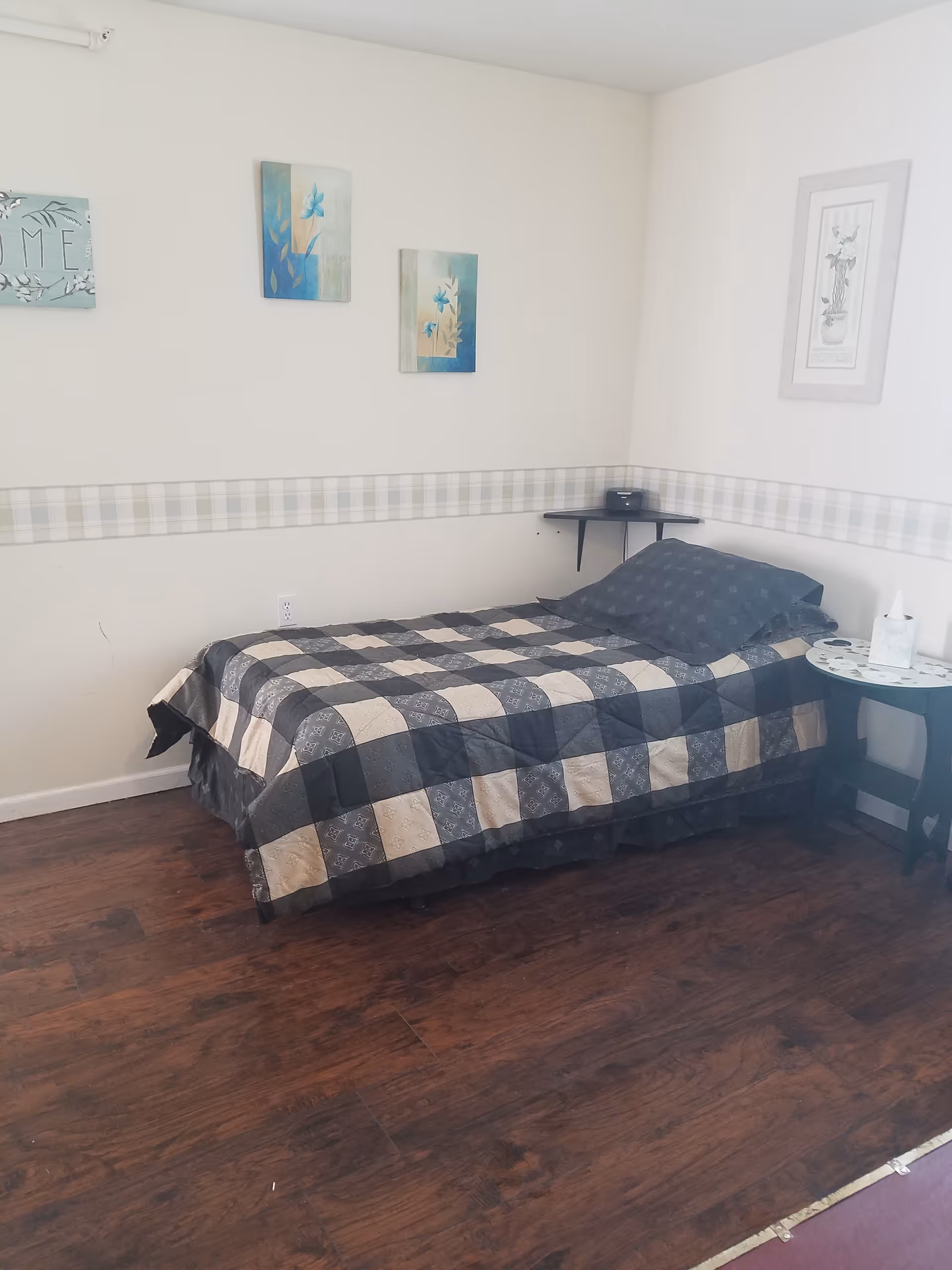 A single bedroom with a plaid quilt, side table, and wall art.