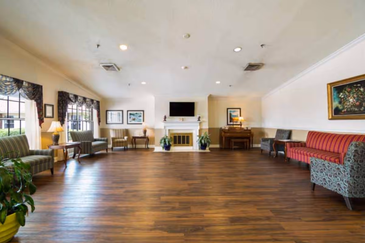 A spacious senior living facility common area with wooden flooring, multiple upholstered chairs and sofas arranged along the walls, large windows with decorative curtains, a fireplace with a TV mounted above it, framed artwork on the walls, and potted plants adding greenery to the room.