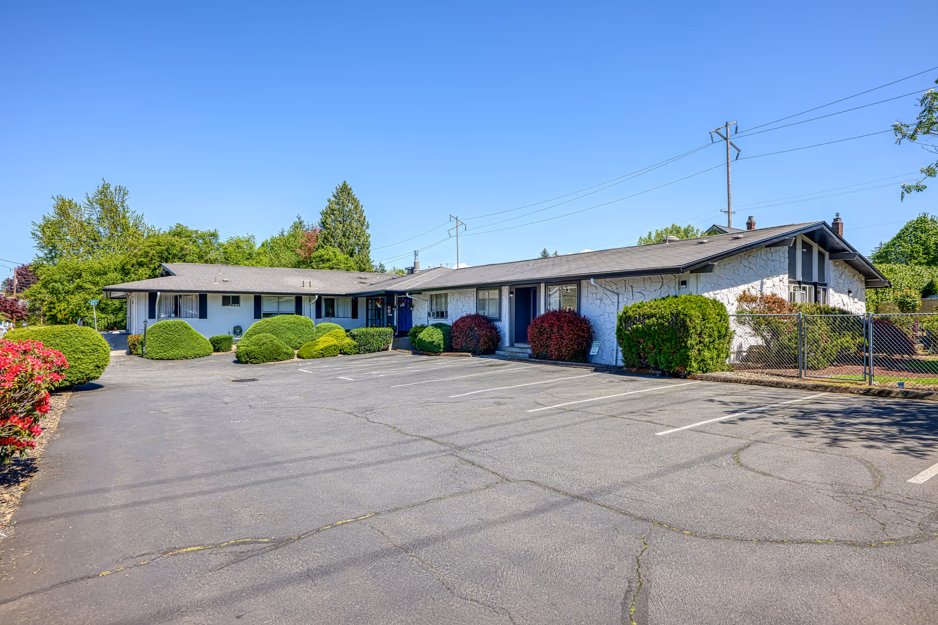 Single-story nursing facility exterior with parking lot, trimmed shrubs, and a chain-link fence.