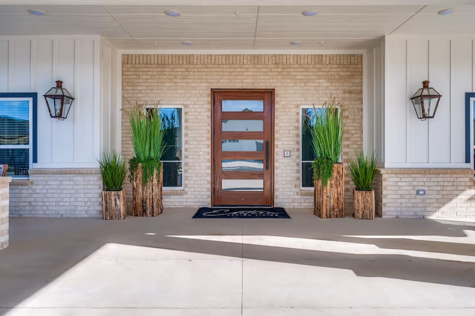 Covered facility entrance with a wooden front door and tall potted plants against a brick wall.
