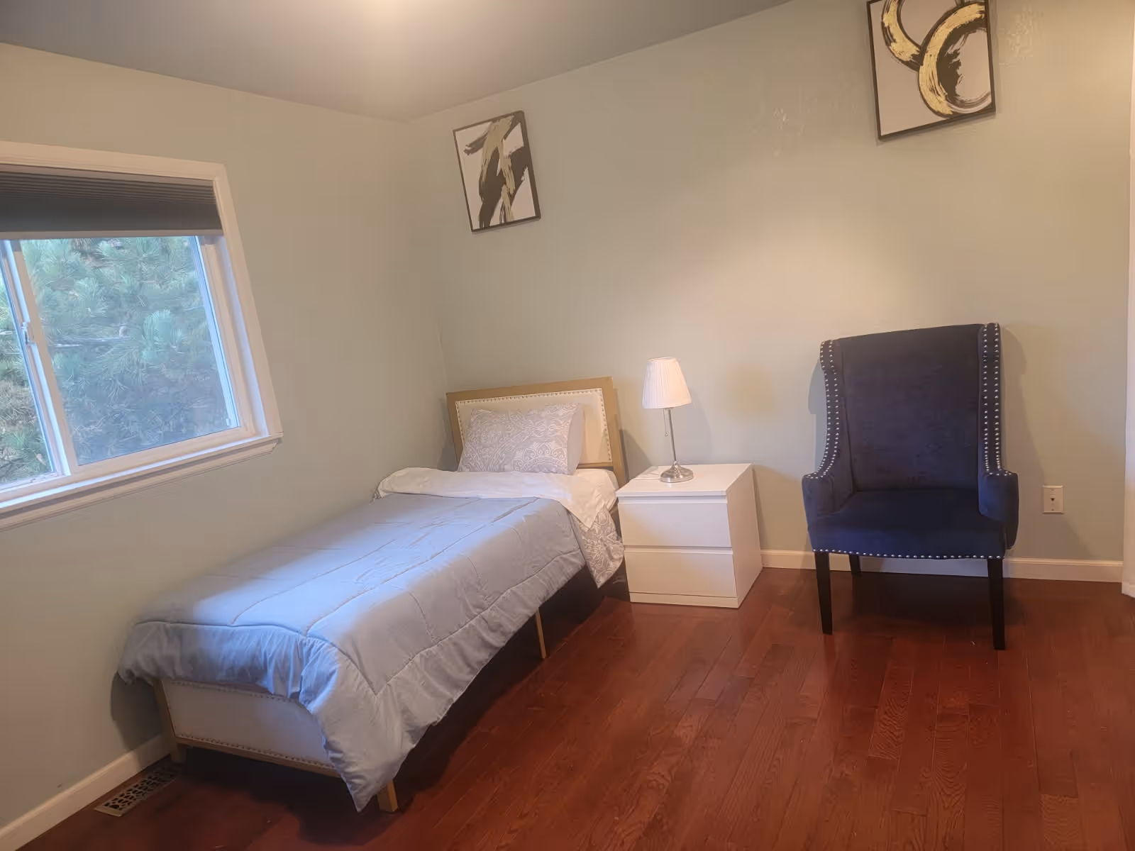 Single bedroom in a senior living facility with bed, nightstand, lamp, and chair.