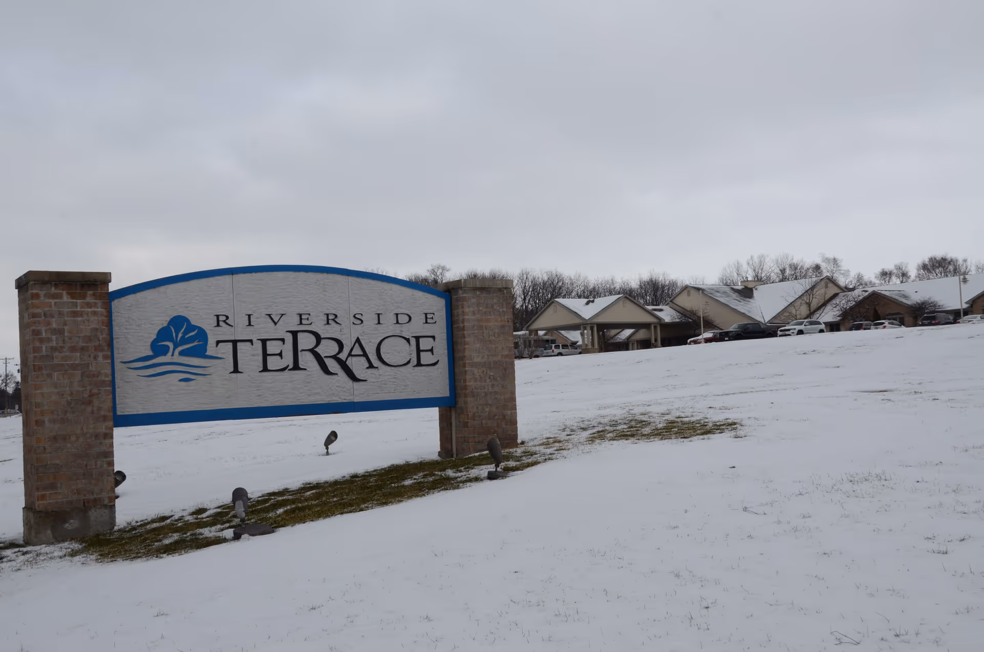 Snowy landscape with Riverside Terrace sign and row of campus-style buildings in distance.