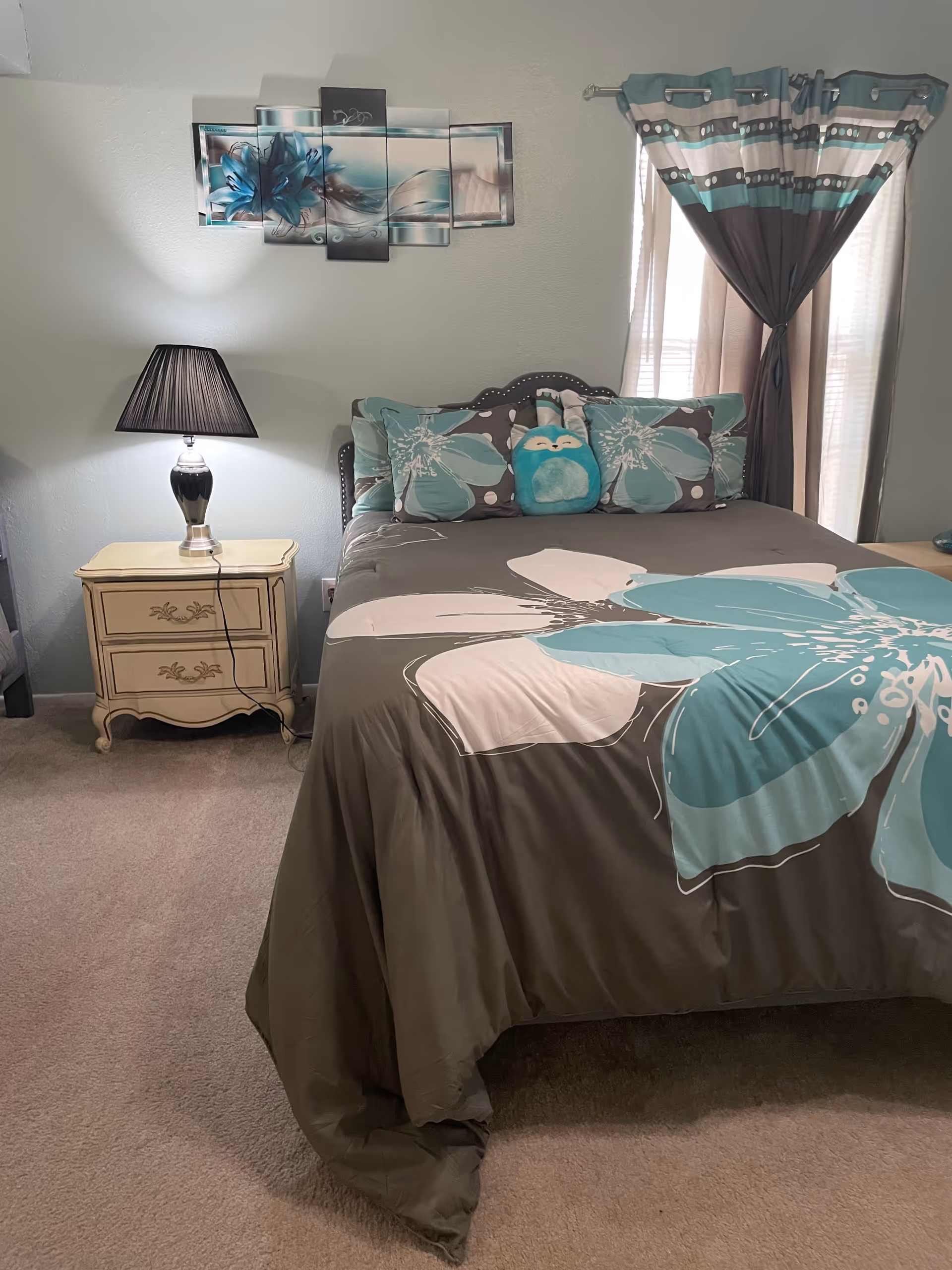 Neatly made bed with teal and gray bedding, nightstand, lamp, and curtains.