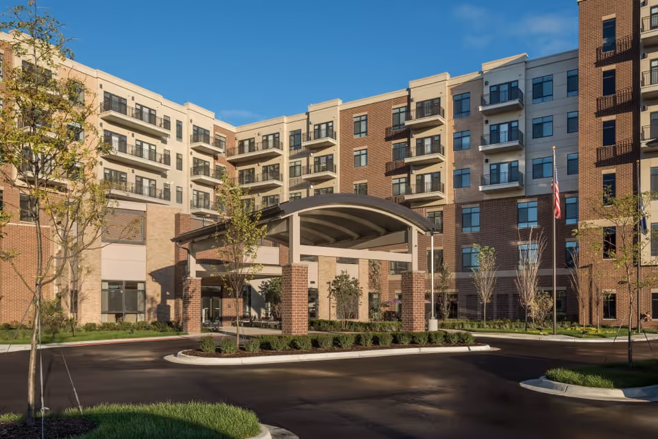 Front entrance of a multi-story senior living building with a covered porte-cochere, balconies, landscaping, and an American flag.