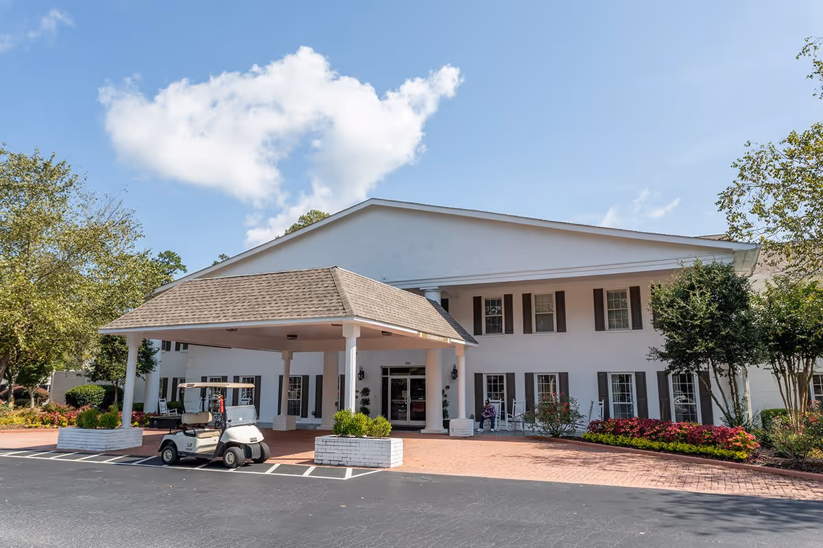 Exterior view of The Retreat at Loganville building with a covered entrance supported by white columns. There is a golf cart parked in front and a person sitting on a rocking chair near the entrance. The area is landscaped with trees, bushes, and flowers under a partly cloudy blue sky.