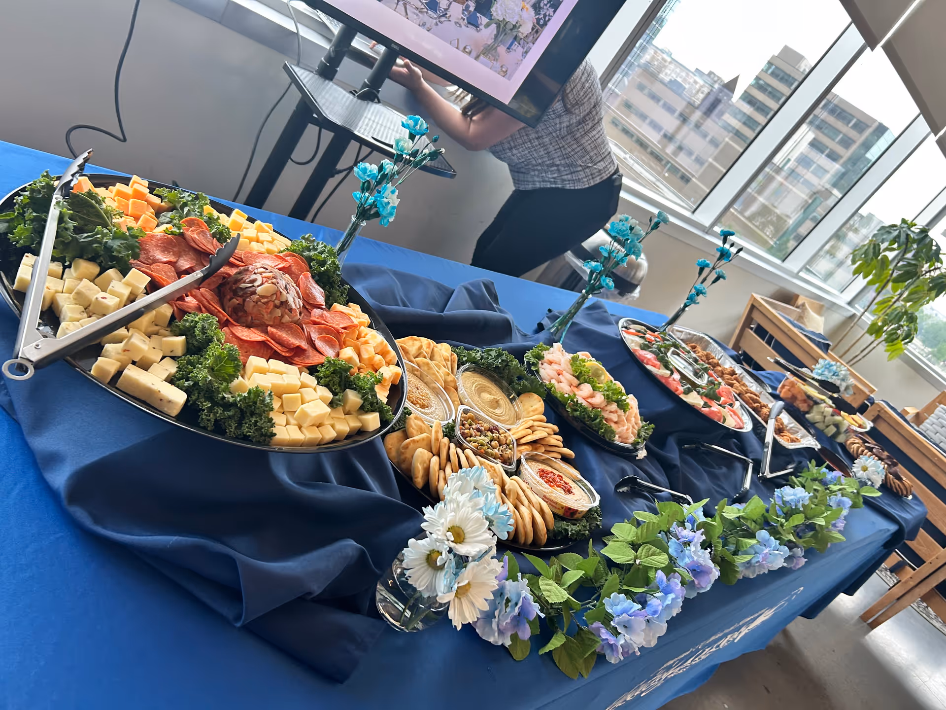 Buffet spread of cheeses, meats, and crackers on a blue tablecloth in a bright dining area.