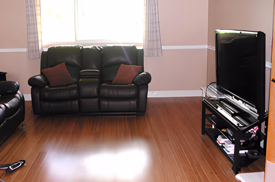 Sunlit living room with a black leather couch, TV on a stand, and wood floors.