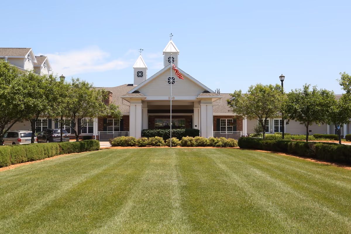 Front exterior view of Melrose Meadows Retirement Community building with a well-maintained lawn, shrubs, trees, and an American flag on a flagpole in the center.