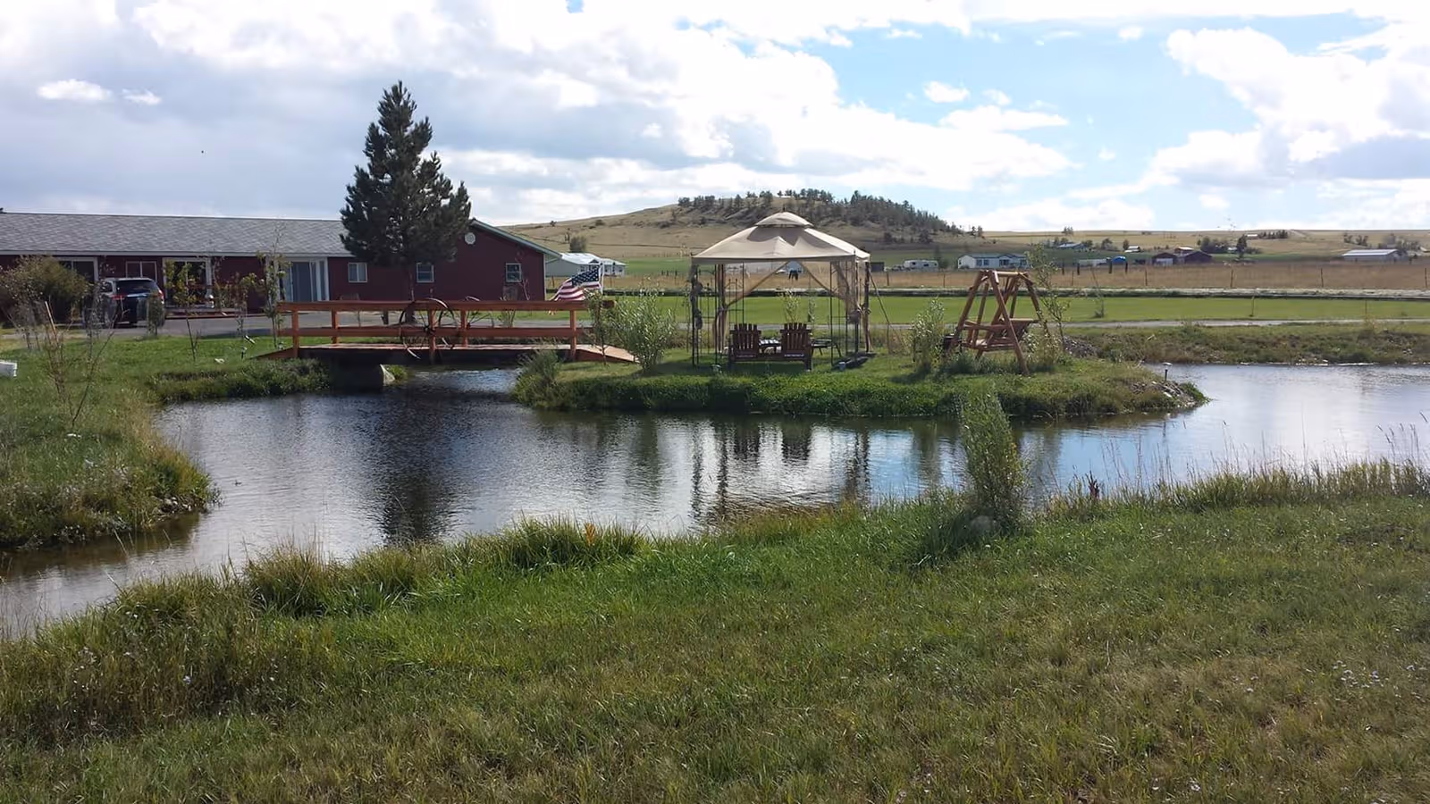 Outdoor pond with wooden deck, gazebo, swings, and red facility building in rural setting.