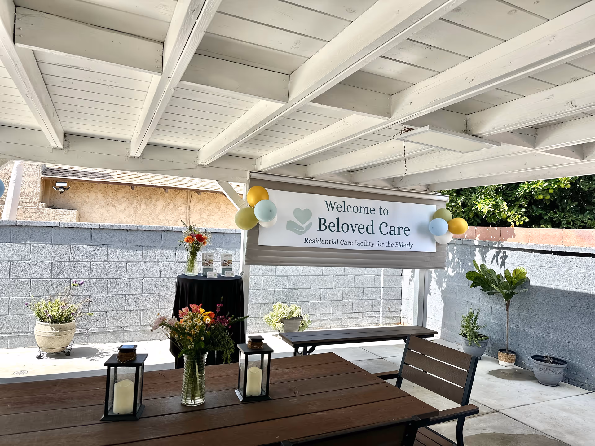 Outdoor covered patio with a Welcome to Beloved Care banner, benches, tables, and potted plants.