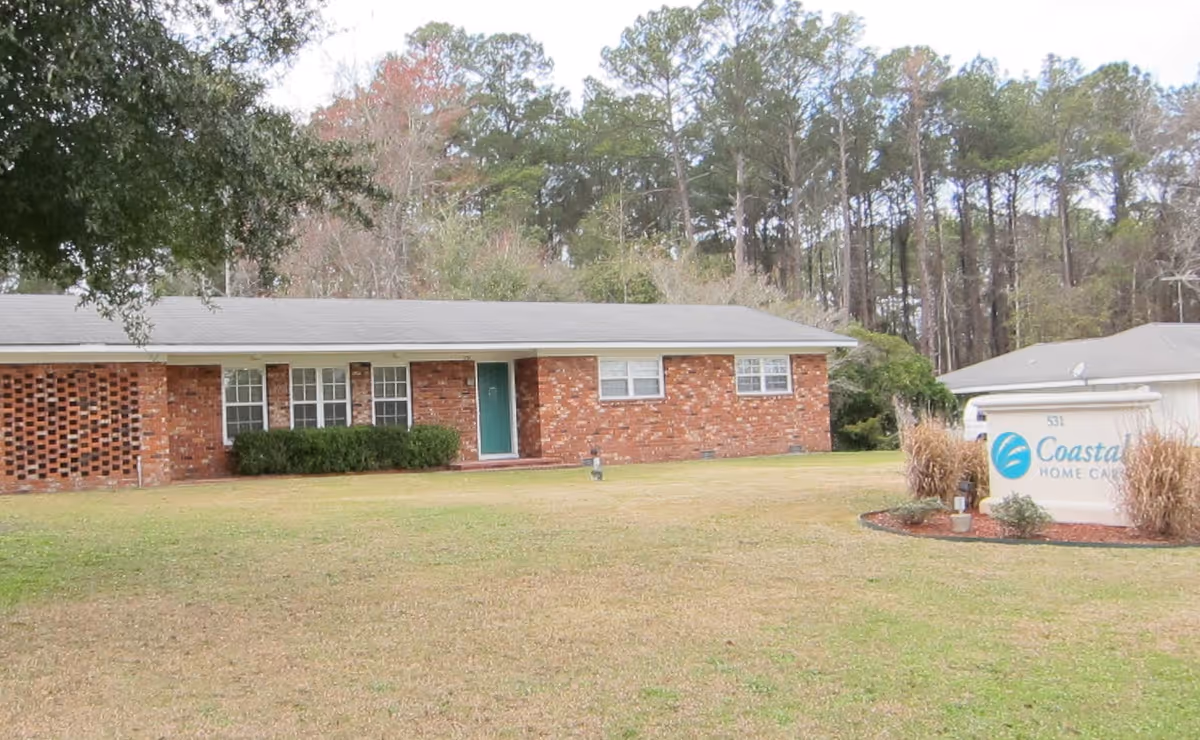 Brick single-story building with green door, lawn foreground, tall trees behind, sign on right.