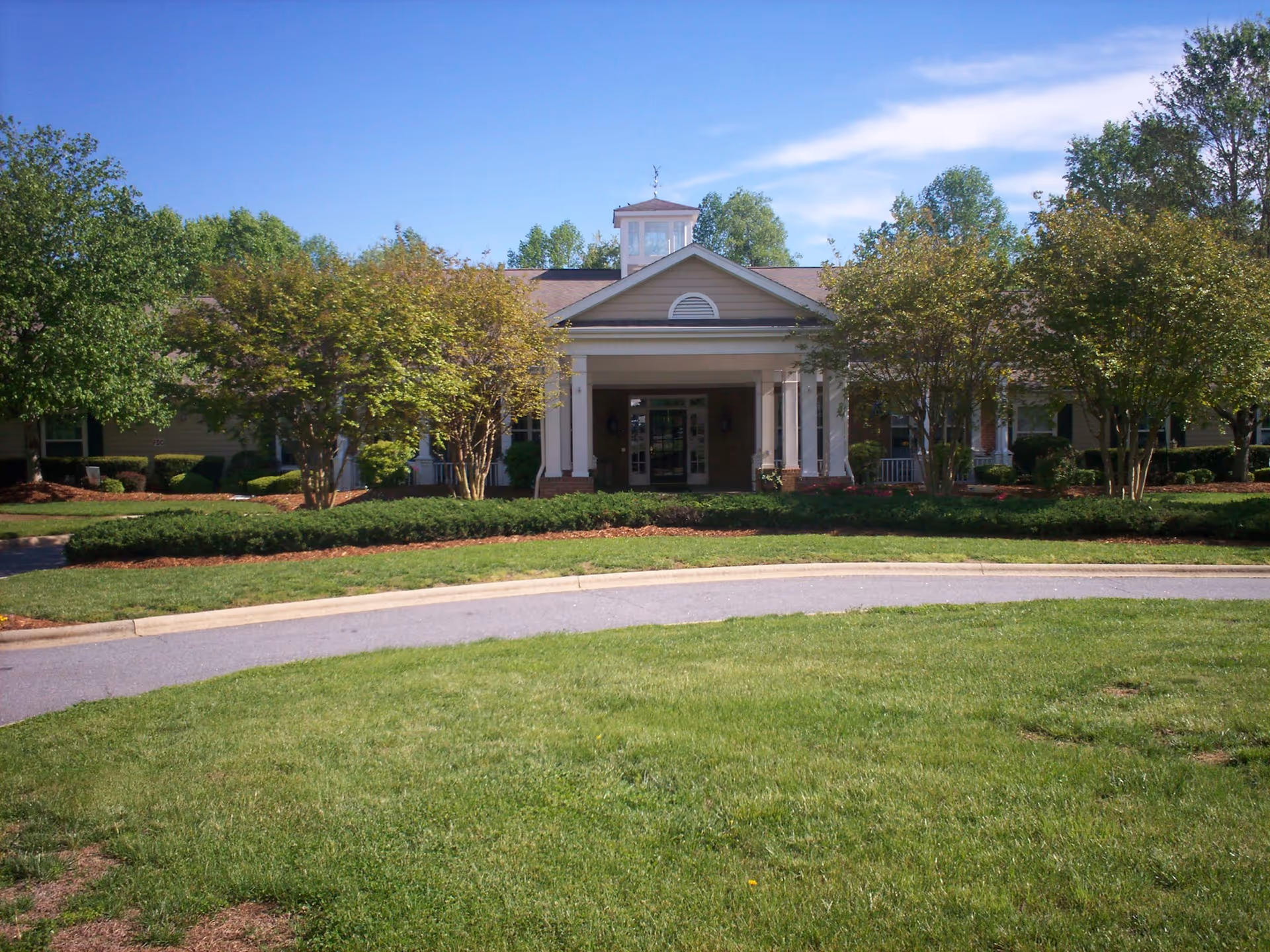 Front entrance of a single-story building with a portico, circular driveway and landscaped lawn and trees.