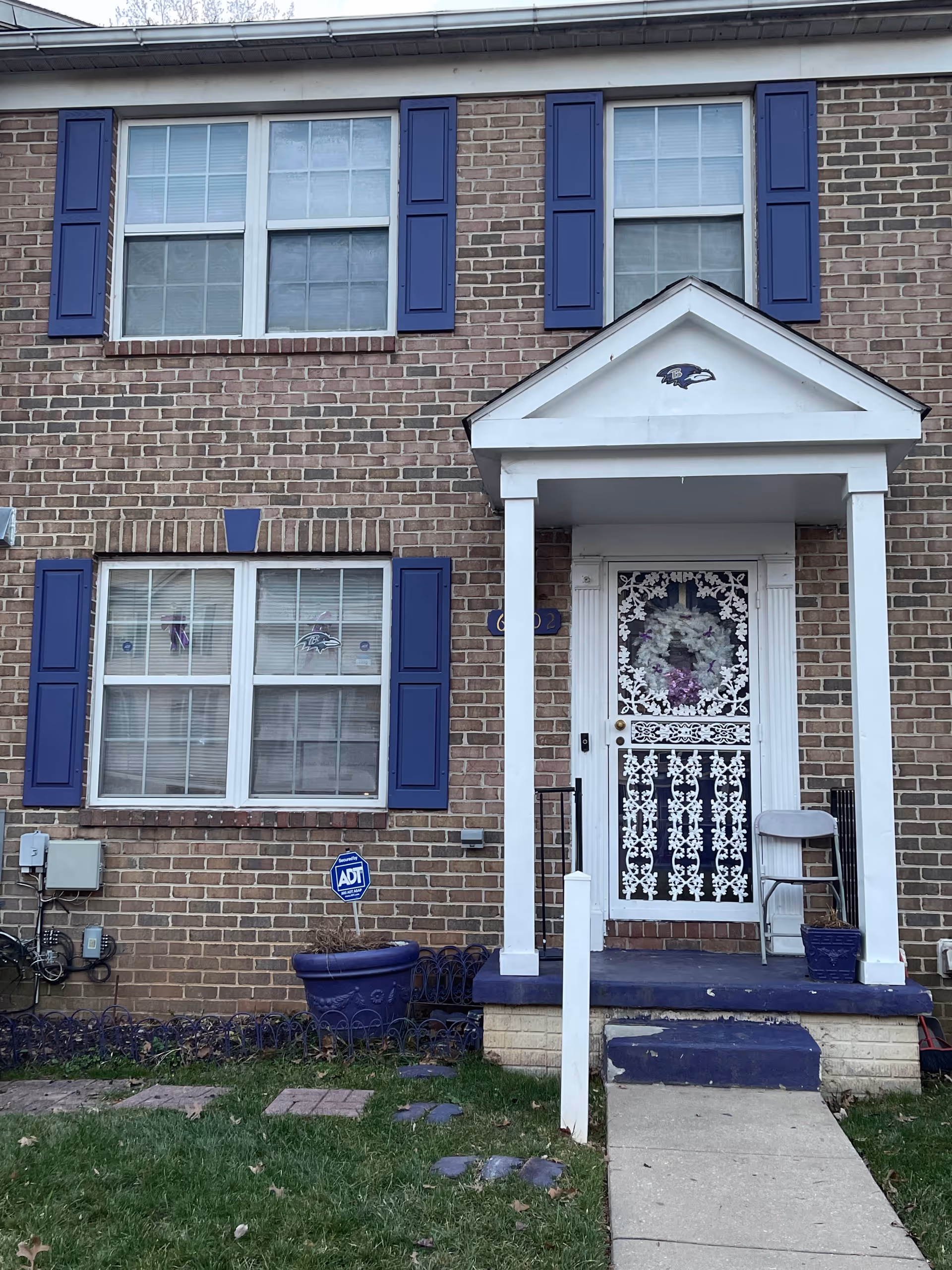 Front of a two-story brick townhouse with blue shutters, a white porch over a decorative front door, and purple-painted steps and planters.