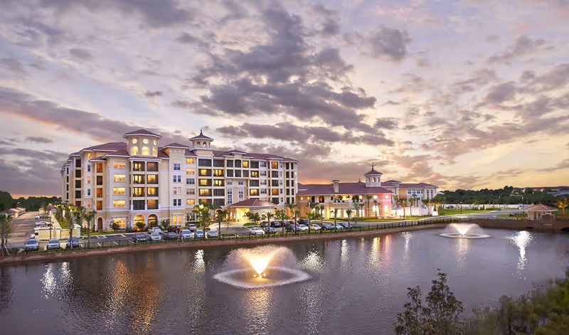 Exterior view of a large multi-story senior living facility named The Terraces at Bonita Springs during sunset, with a pond featuring two illuminated fountains in the foreground and a parking lot with cars along the building.