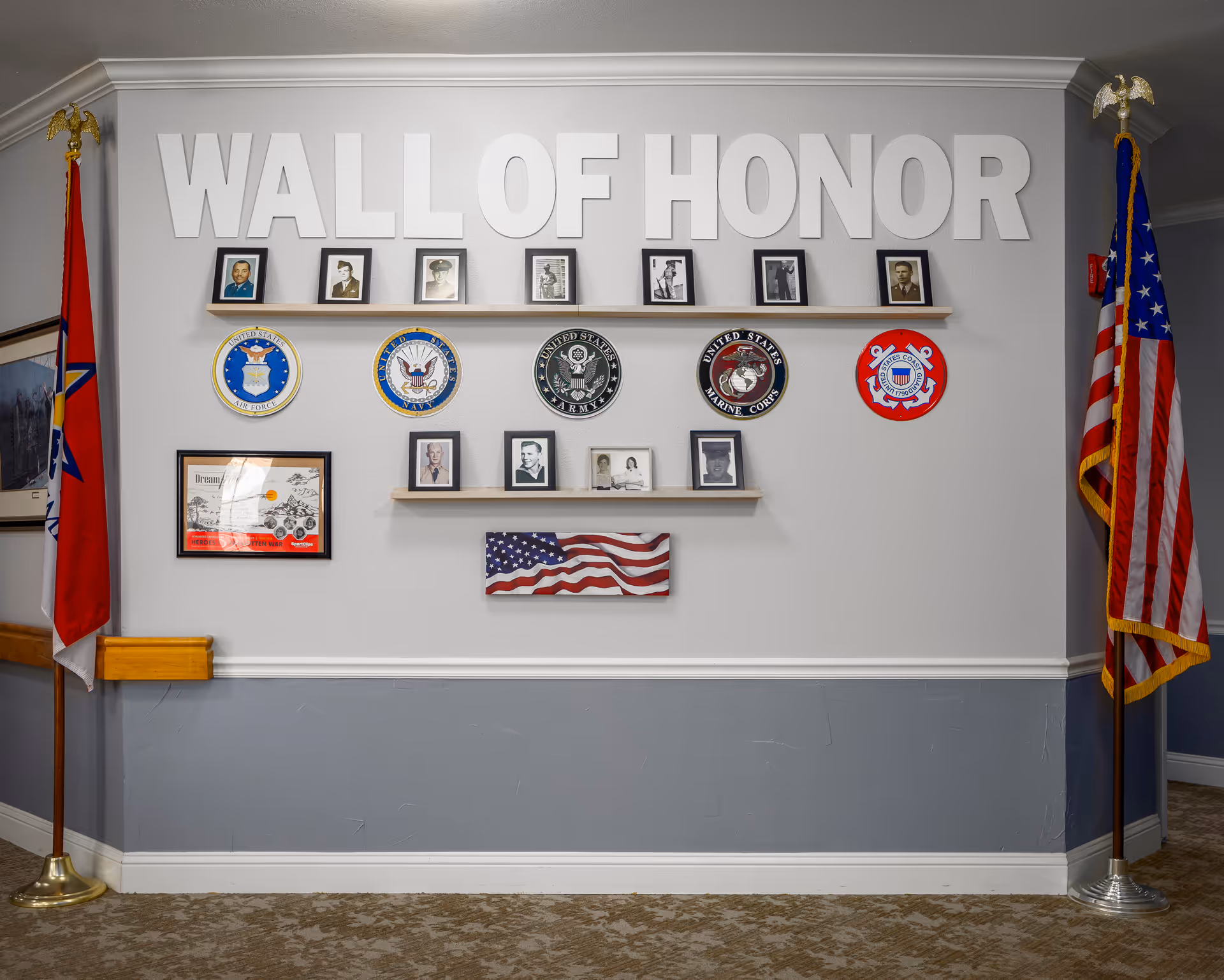 A Wall of Honor display featuring framed photographs of veterans, military branch emblems including Air Force, Navy, Army, Marine Corps, and Coast Guard, an American flag artwork, and two flags on stands on either side of the wall.