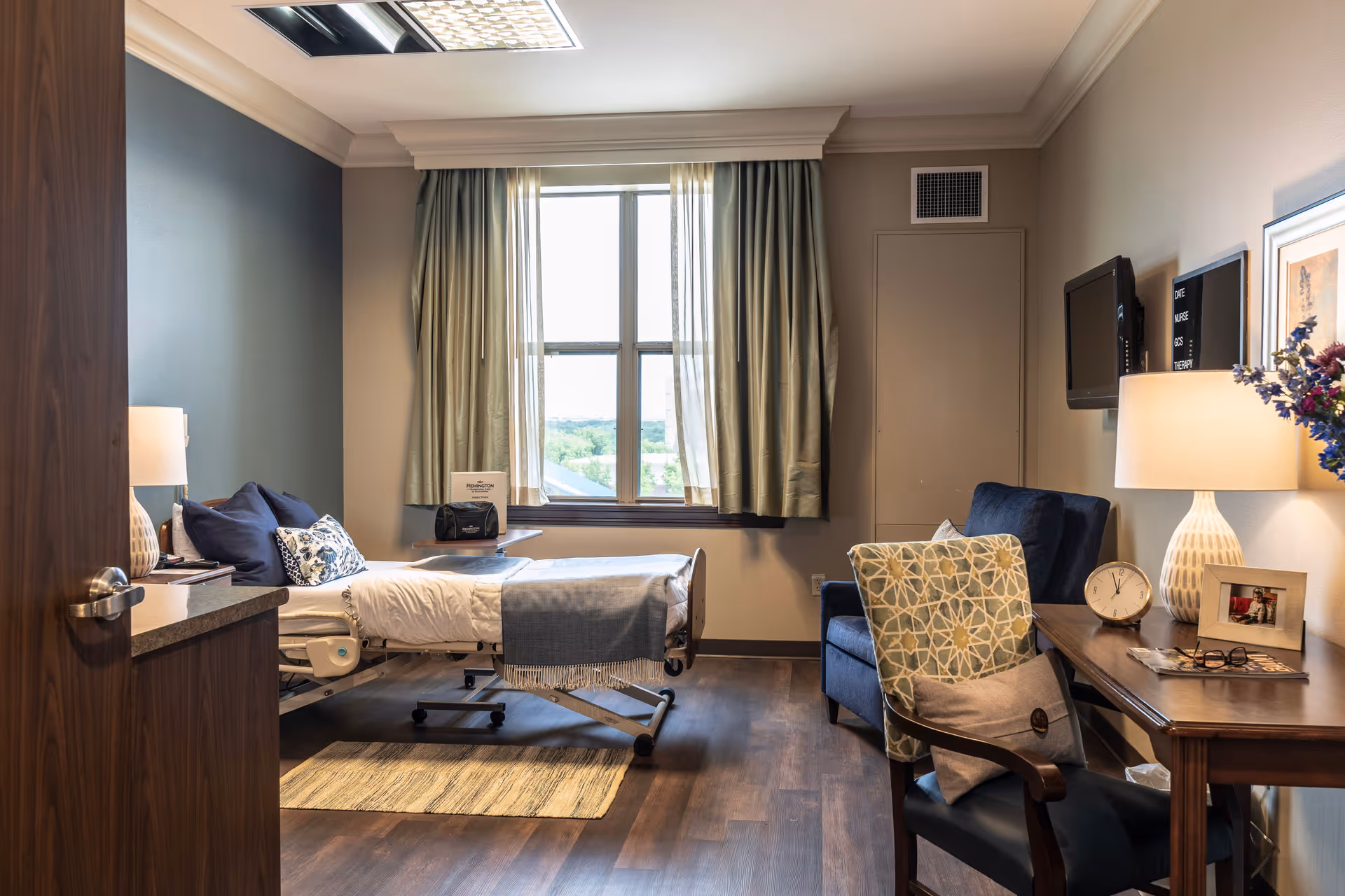 A well-lit hospital-style bedroom in a senior care facility with bed, desk, and seating near window.