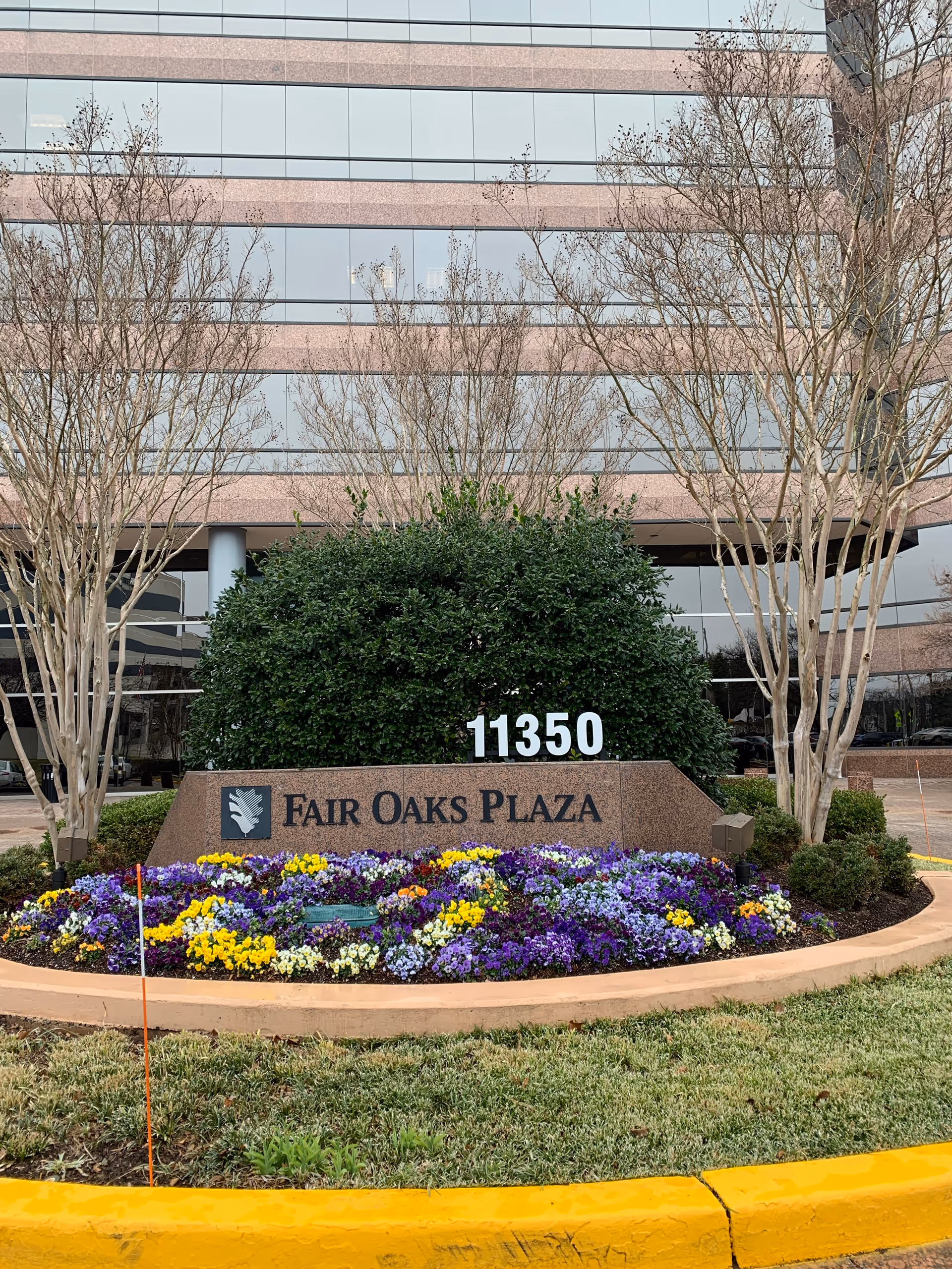 Exterior building front with landscaped sign reading 11350 Fair Oaks Plaza and colorful flowers.