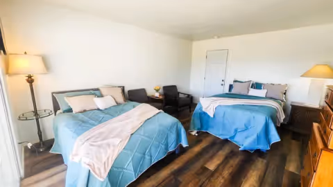 Bright bedroom with two double beds dressed in blue bedding, nightstands with lamps, chairs and wood-look flooring.