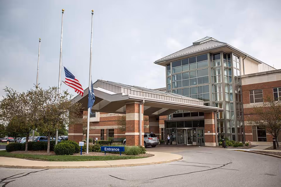 Front entrance of a brick-and-glass senior living building with a covered drop-off, flagpoles and an "Entrance" sign.