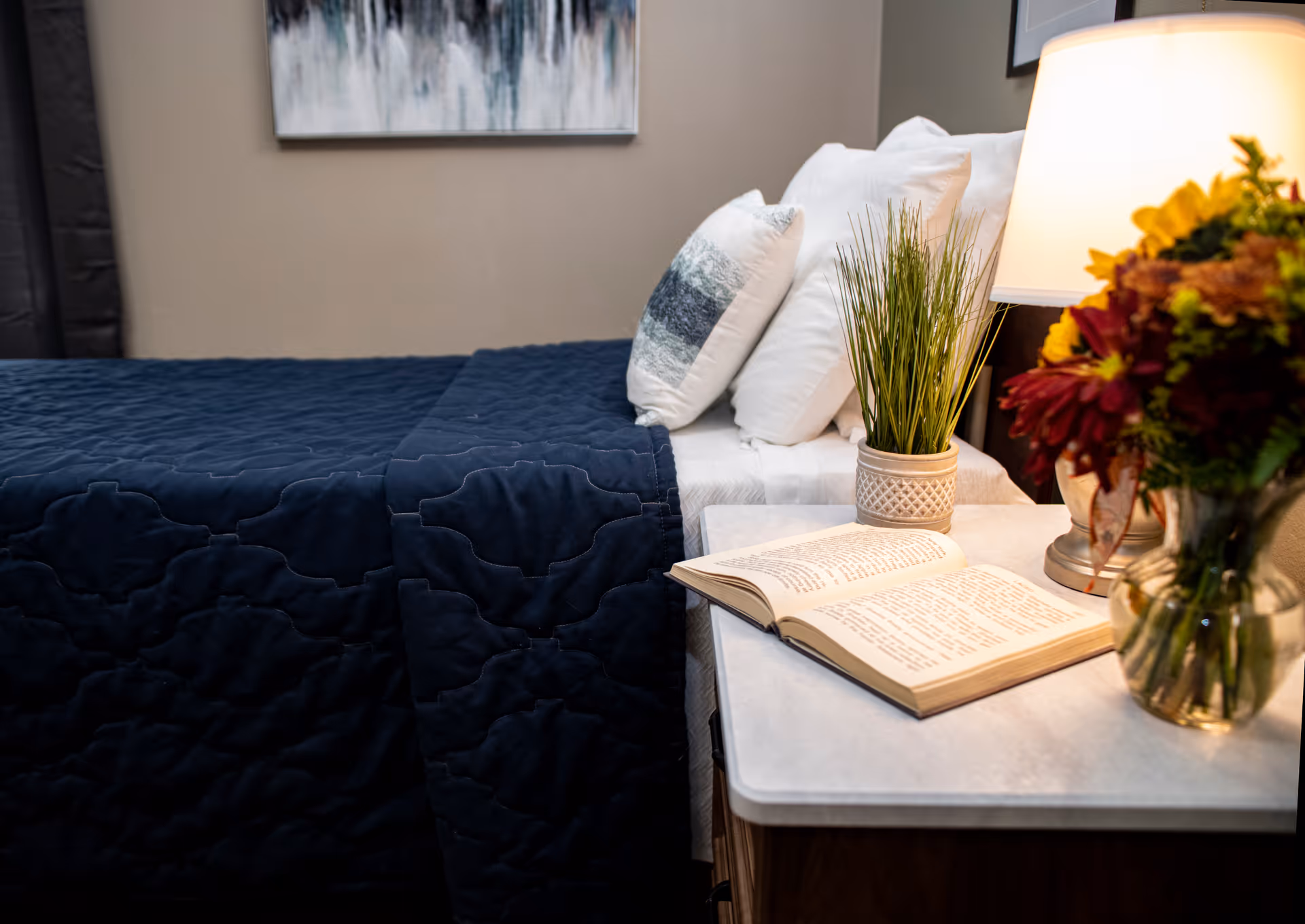 A bedside table with an open book, lamp, potted plant and flowers next to a neatly made bed with a dark blue quilt and pillows.