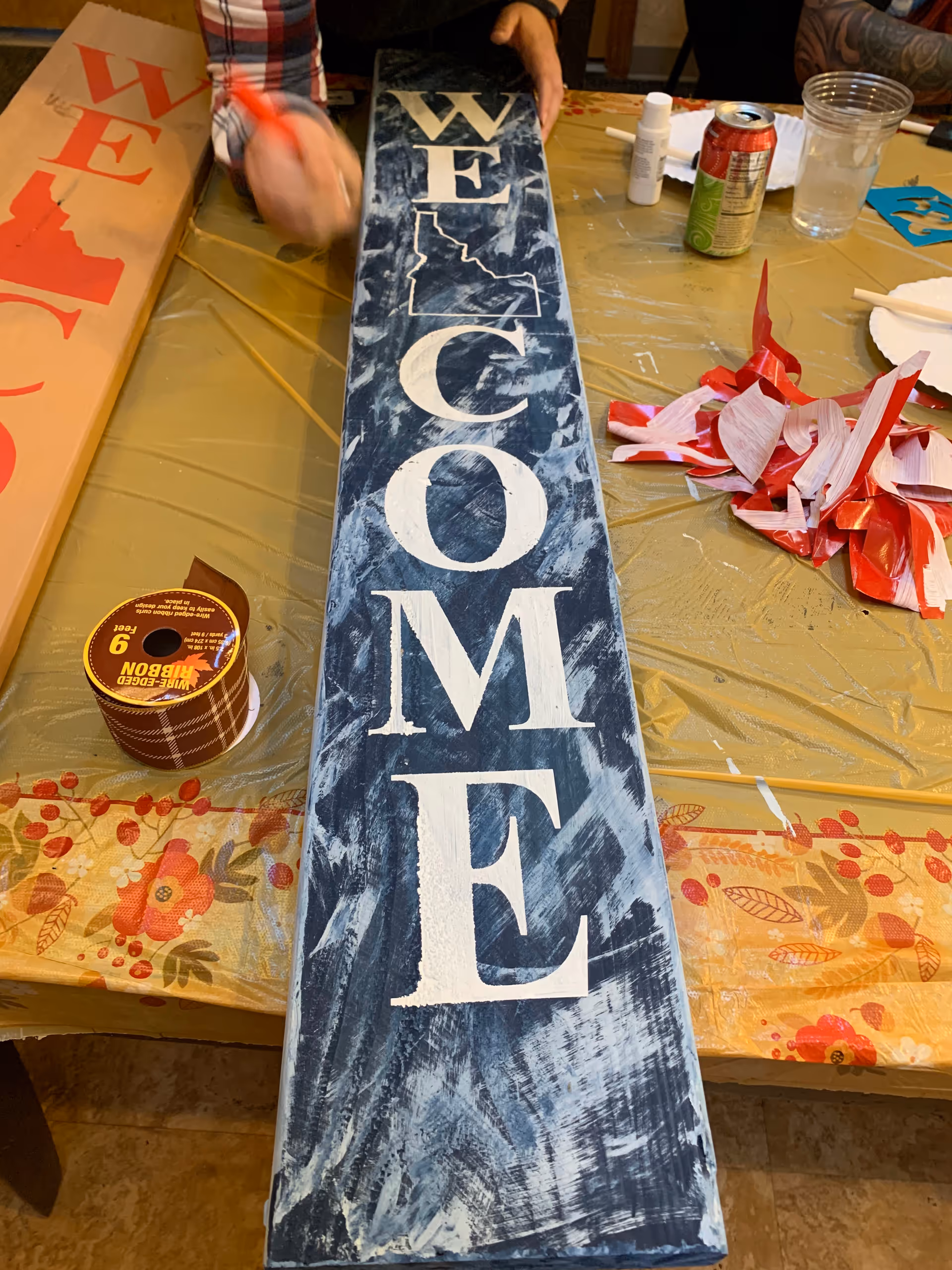 A hand paints a blue distressed 'WELCOME' sign on a craft table.